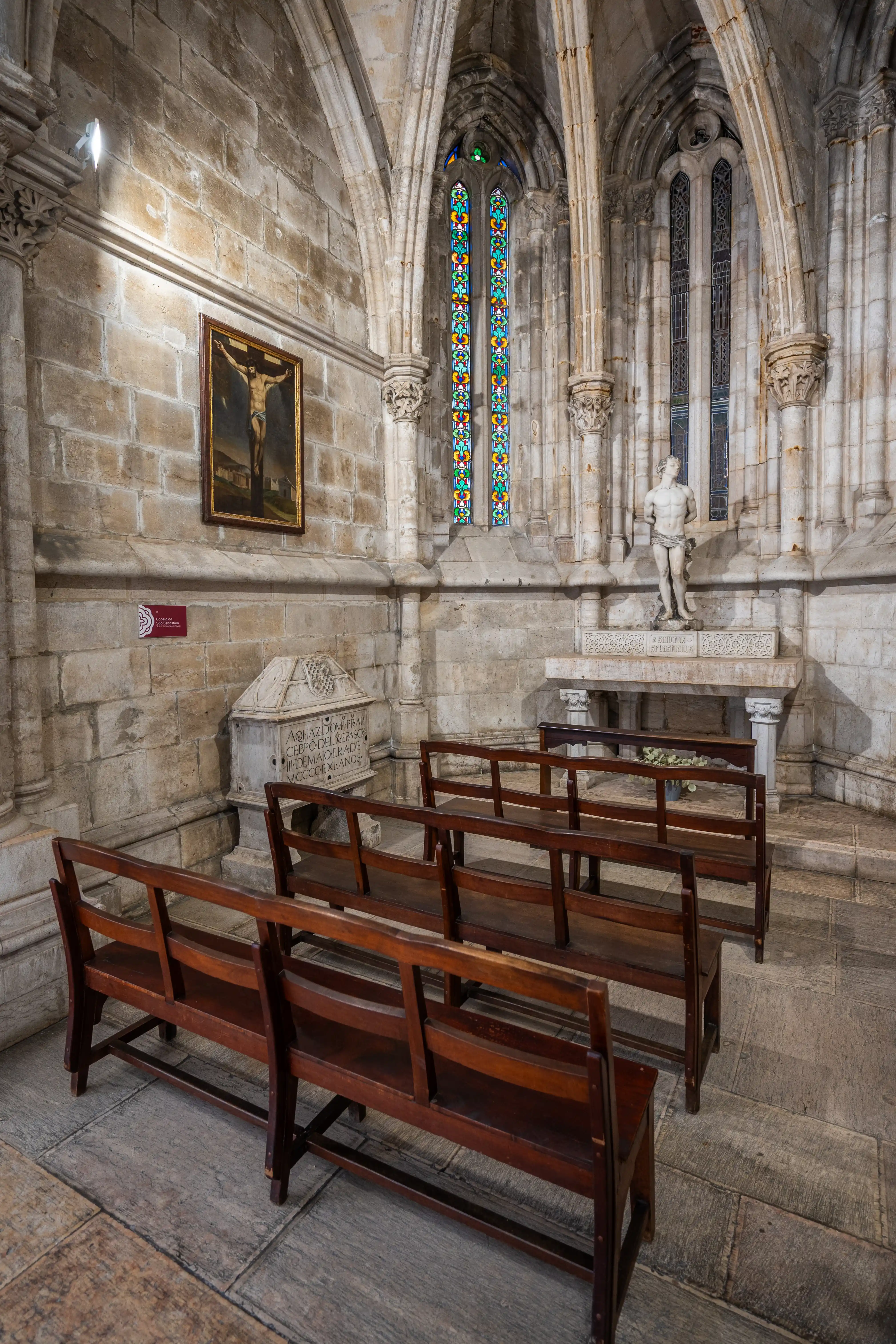 Interior of a historic chapel showing wooden pews, a painting of the Crucifixion, a sarcophagus, a marble statue, and stained-glass windows.