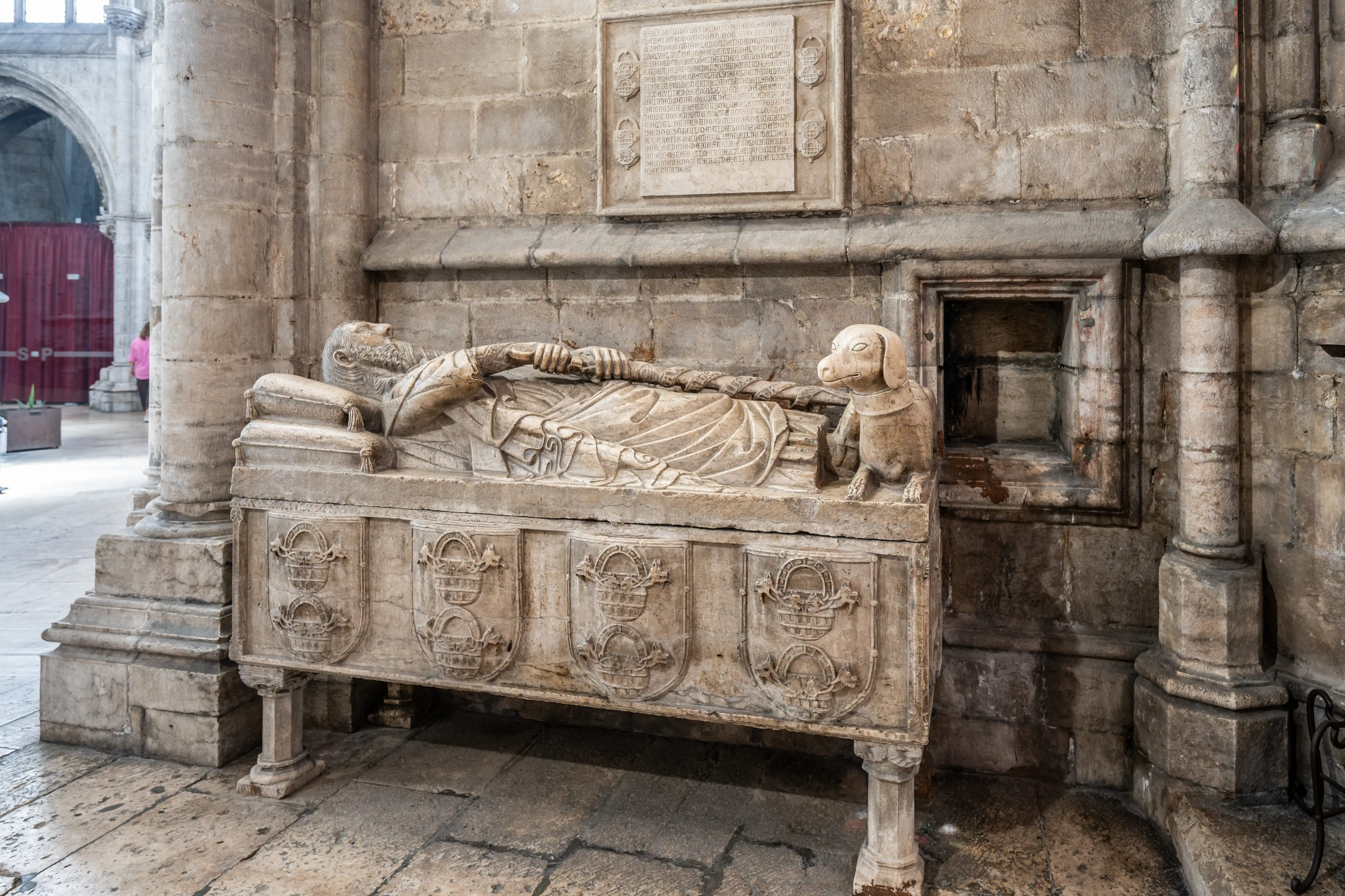 A stone sarcophagus with the effigy of a bearded man and a dog sculpture at his feet, set against a stone wall with a memorial plaque in a grand historical building.