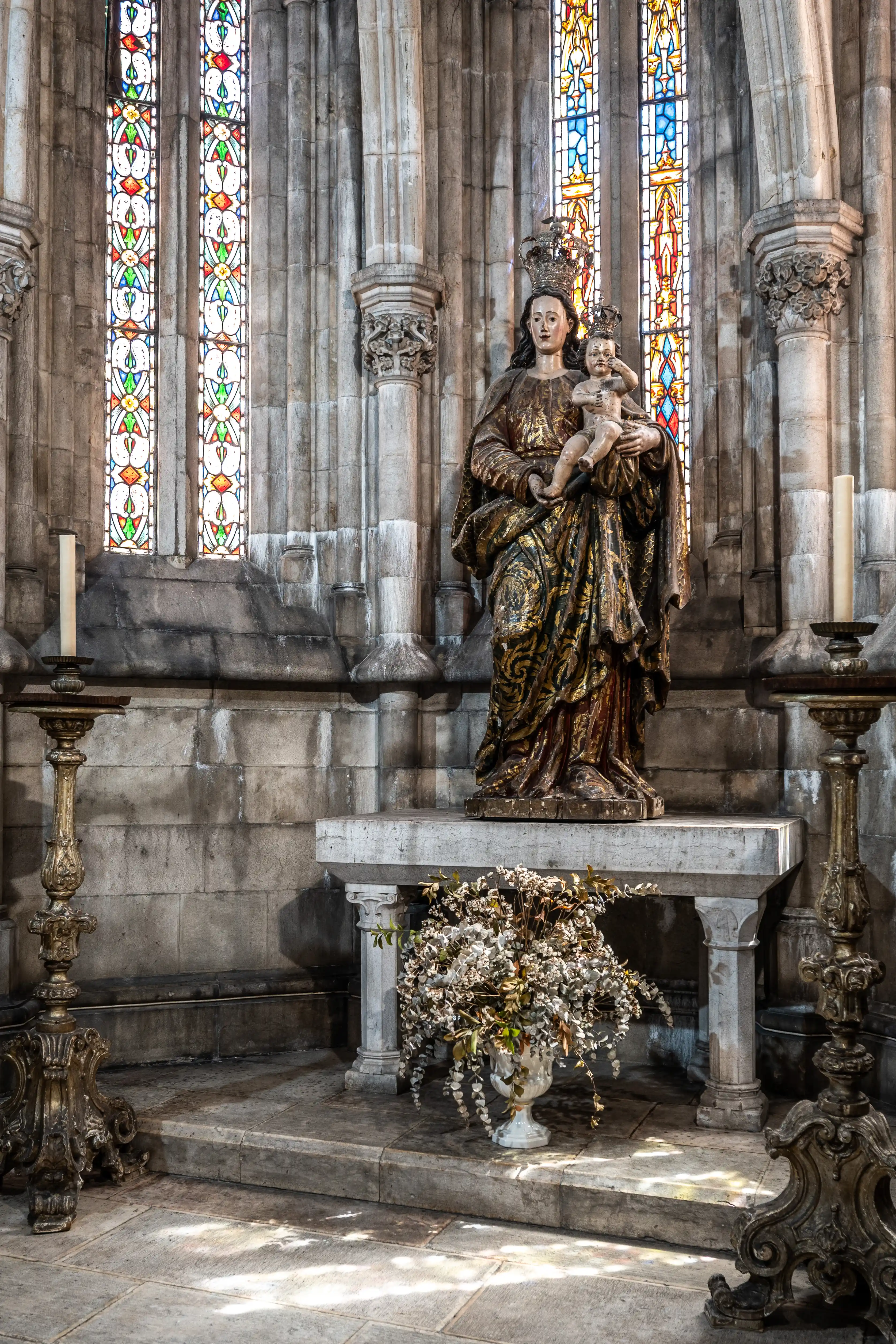  A religious statue of the Virgin Mary holding baby Jesus, crowned and dressed in a golden, ornate robe, stands on a stone altar. Behind the statue are tall stone columns and brightly coloured stained glass windows. Two decorative candelabras, one with a lit candle, and a vase of dried flowers are also visible.