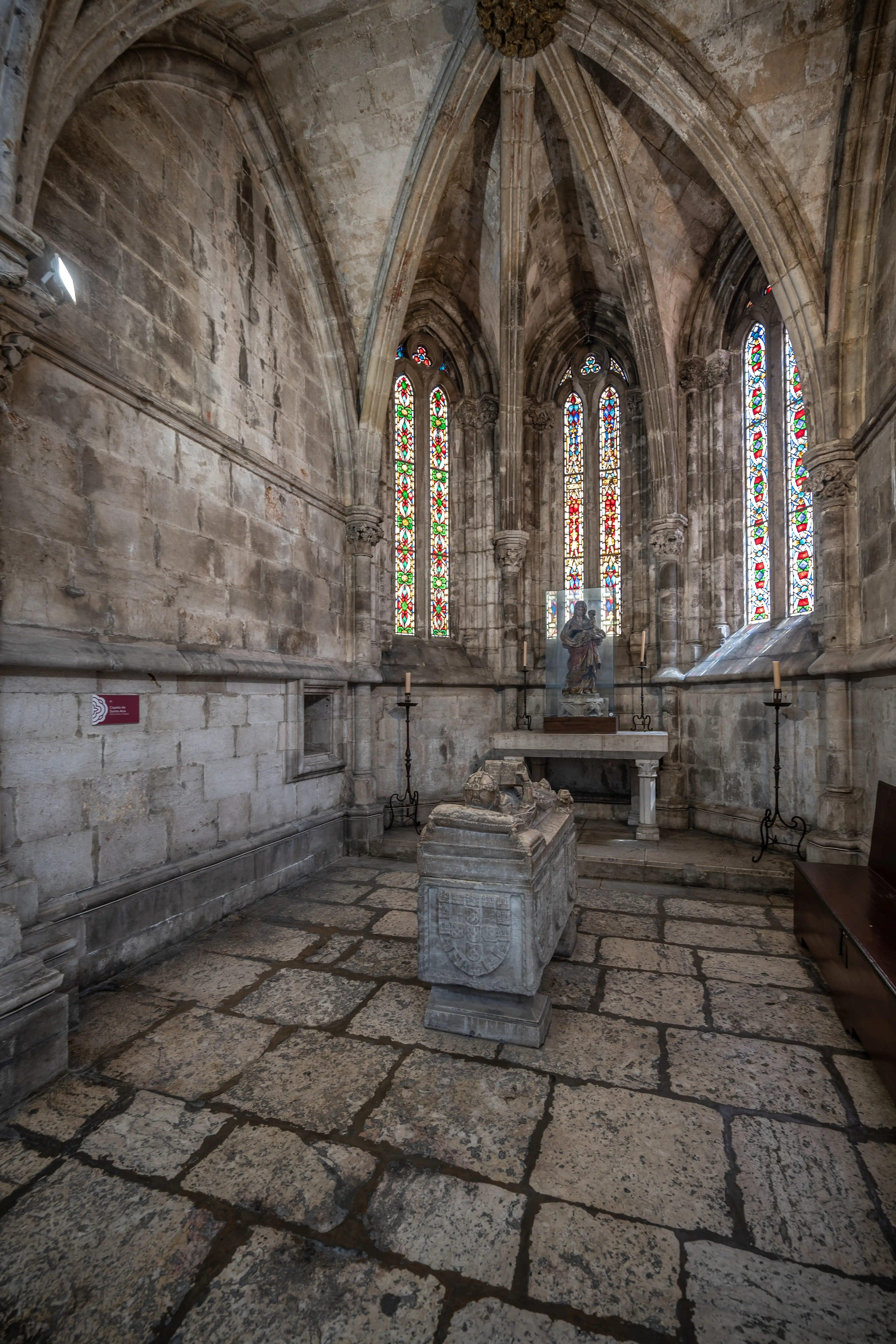 Interior view of a historic chapel featuring a stone sarcophagus in the foreground, colourful stained glass windows, vaulted stone ceilings, and stone block walls.