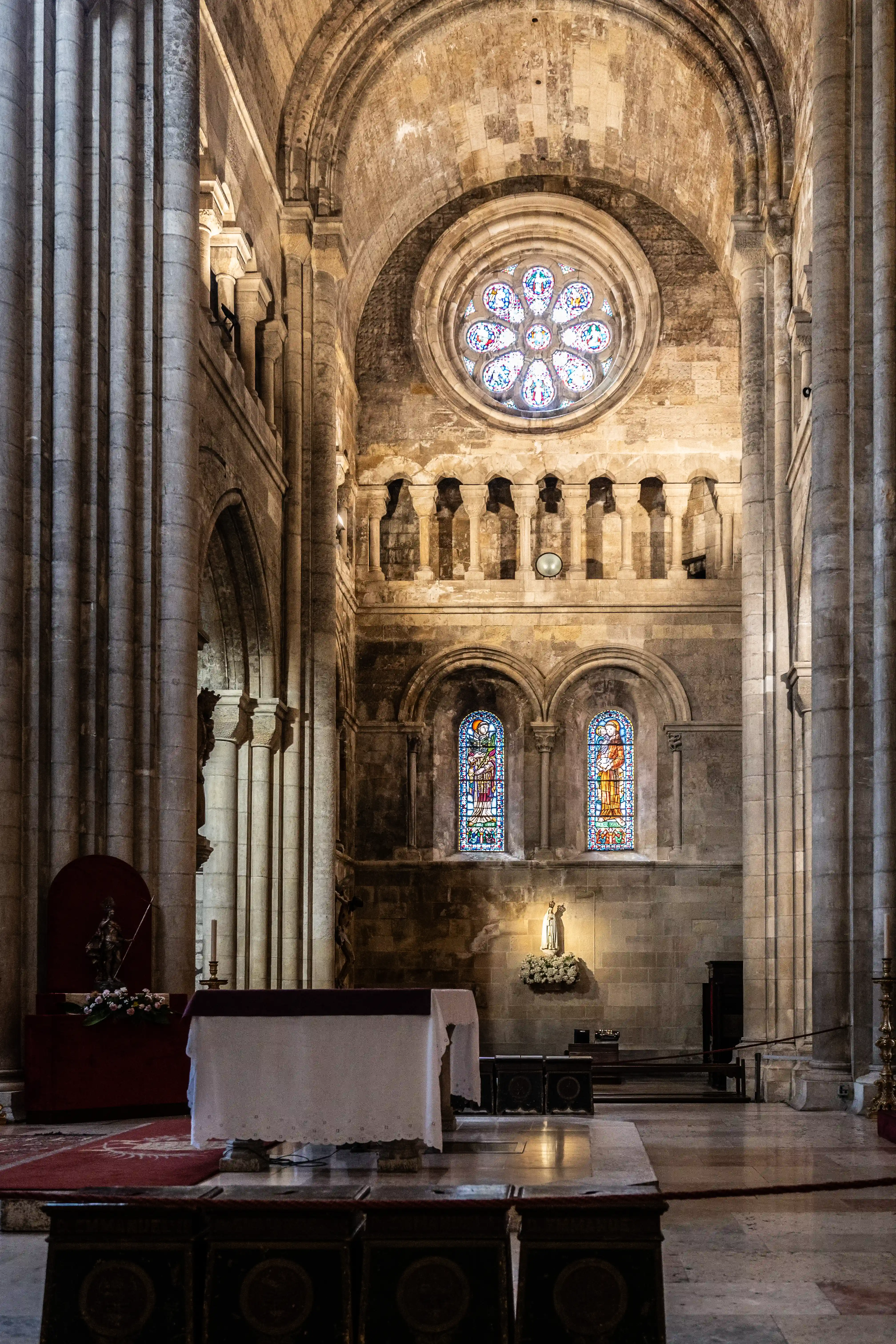 Inside of a cathedral with large stone columns, a rose stained-glass window, two rectangular stained-glass windows, an altar, and a statue of a religious figure.