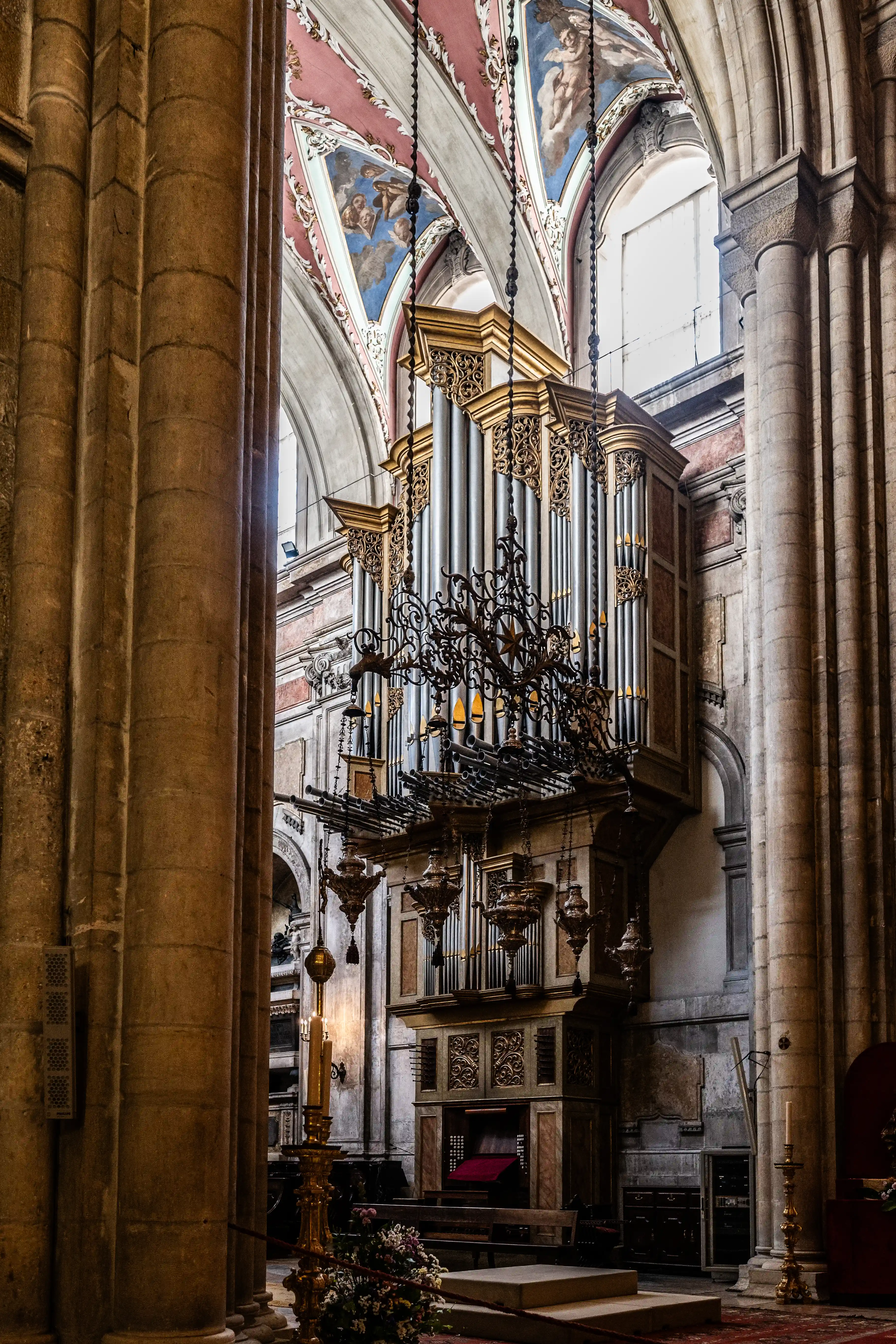 A large, ornate pipe organ inside a cathedral, with detailed silver pipes and gold decorative elements. The vaulted ceiling above features painted frescoes of angels. Large stone columns line the sides of the image, and a lit candelabra stands in the foreground.