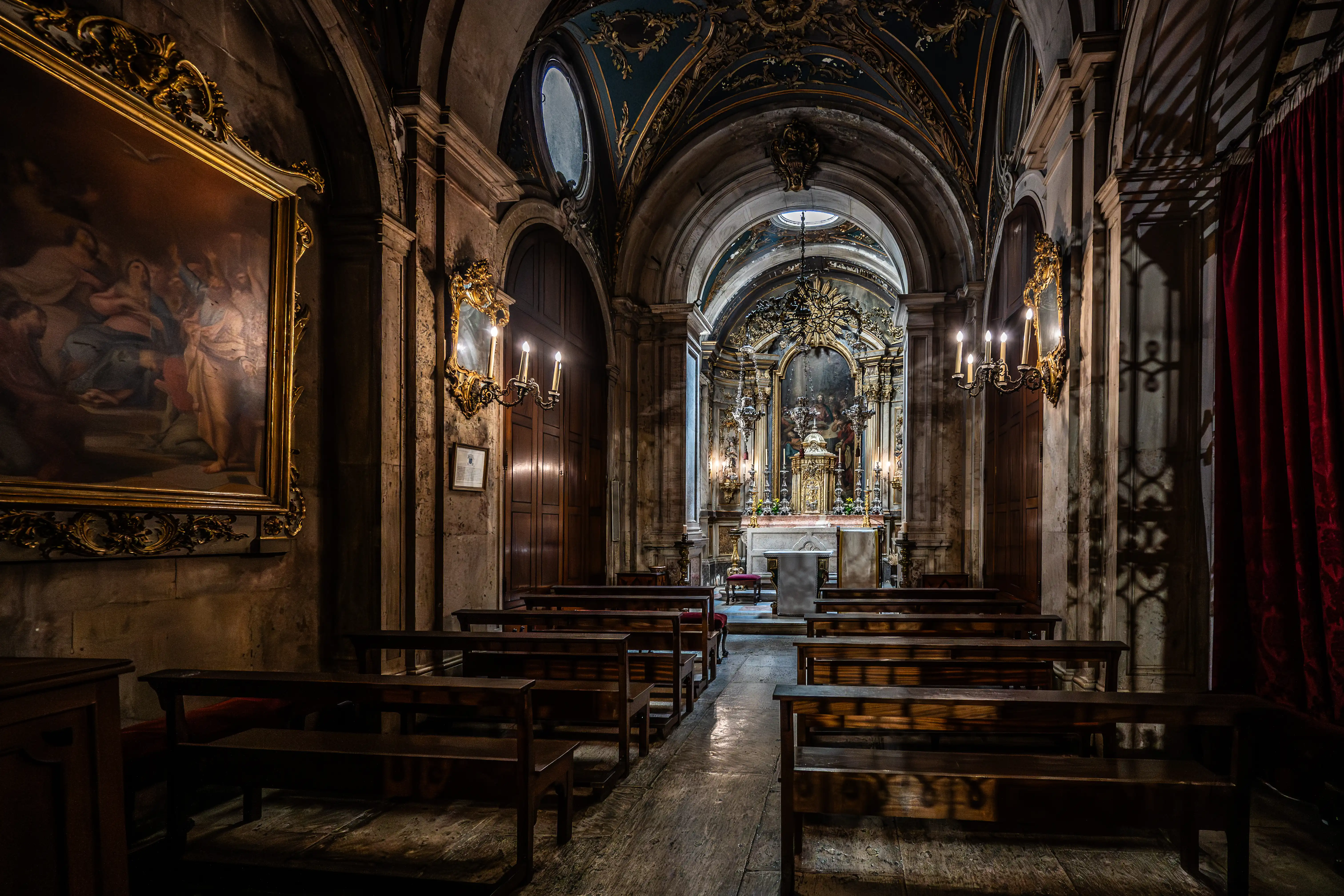 Interior of a dimly lit, ornate chapel with rows of wooden pews leading towards a decorated altar. A large framed painting hangs on the left wall, and candelabra illuminate the space. A red curtain is visible on the right.