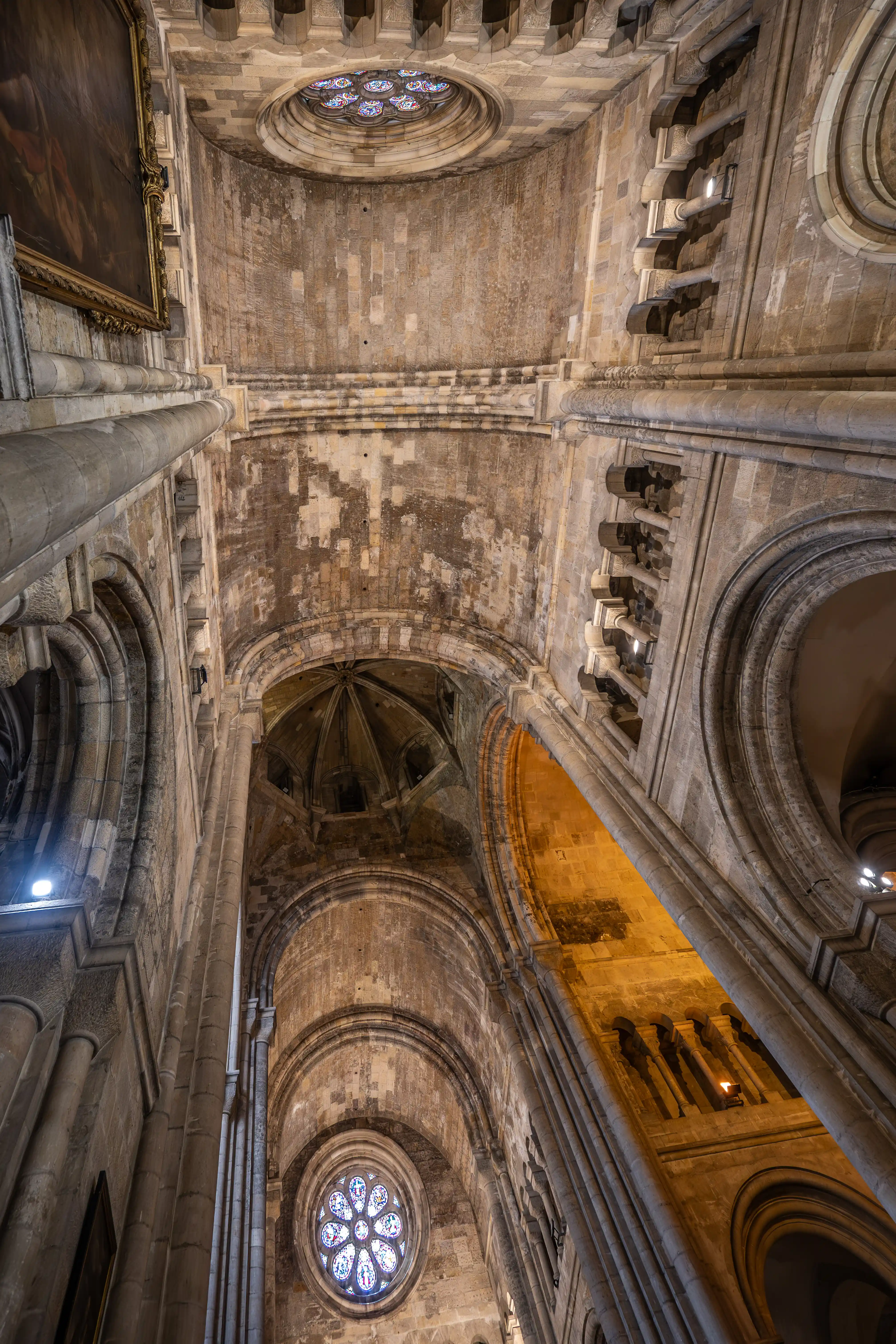 A wide-angle, low-angle view upwards into the interior of a grand cathedral, showcasing its impressive stone architecture, vaulted ceilings, and two circular rose windows filled with colourful stained glass.