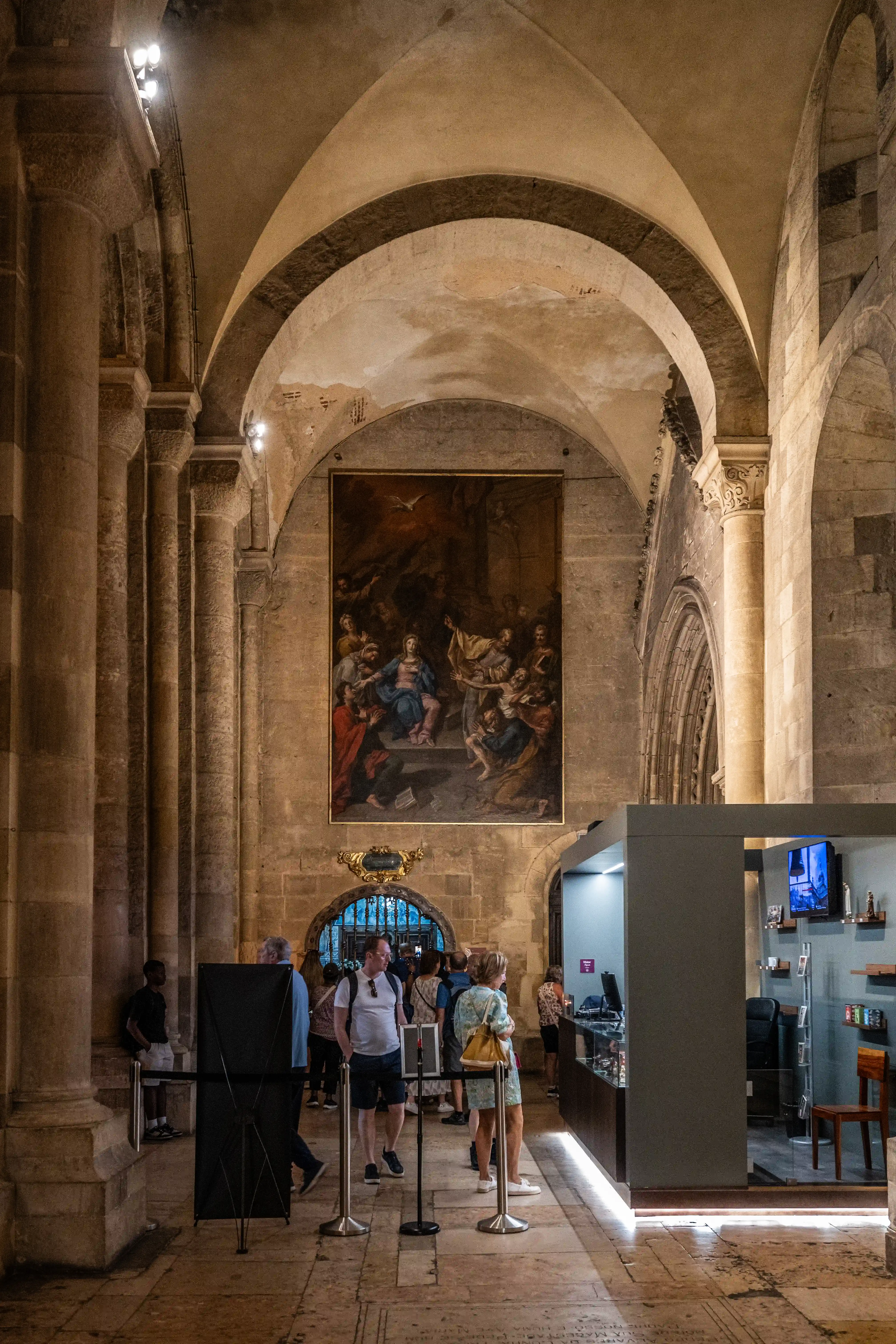 A wide-angle interior shot of a historic building with stone columns and arched ceilings. A large painting hangs on the far wall. Visitors are seen walking through the space, and a modern exhibition area is visible on the right.