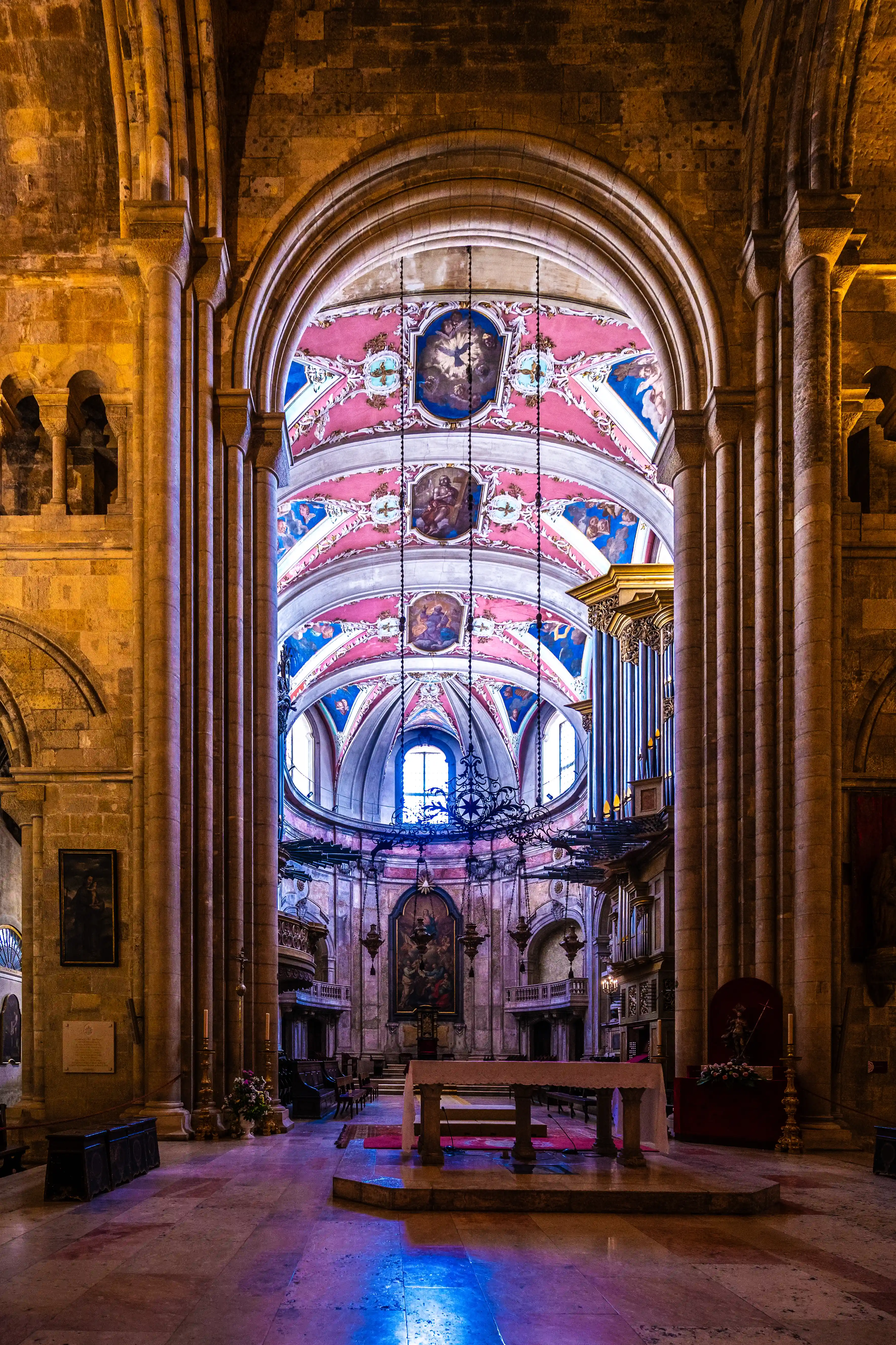 Interior view of a cathedral showcasing a richly decorated vaulted ceiling with religious paintings, stone columns, a pipe organ, and an altar illuminated by blue light