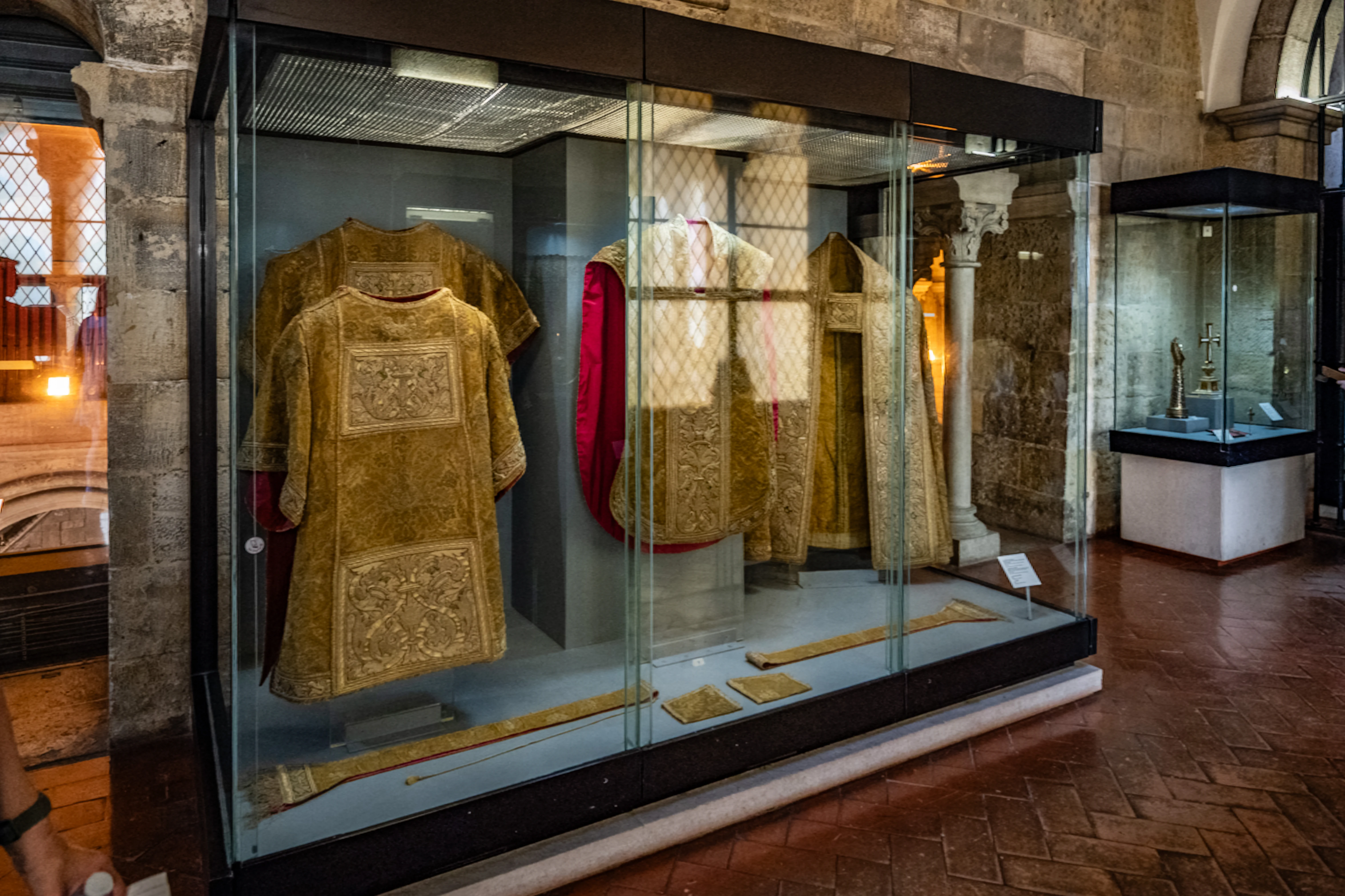 Several gold-coloured embroidered ecclesiastical robes are displayed in a glass case, with further religious artefacts visible in a smaller case in the background.