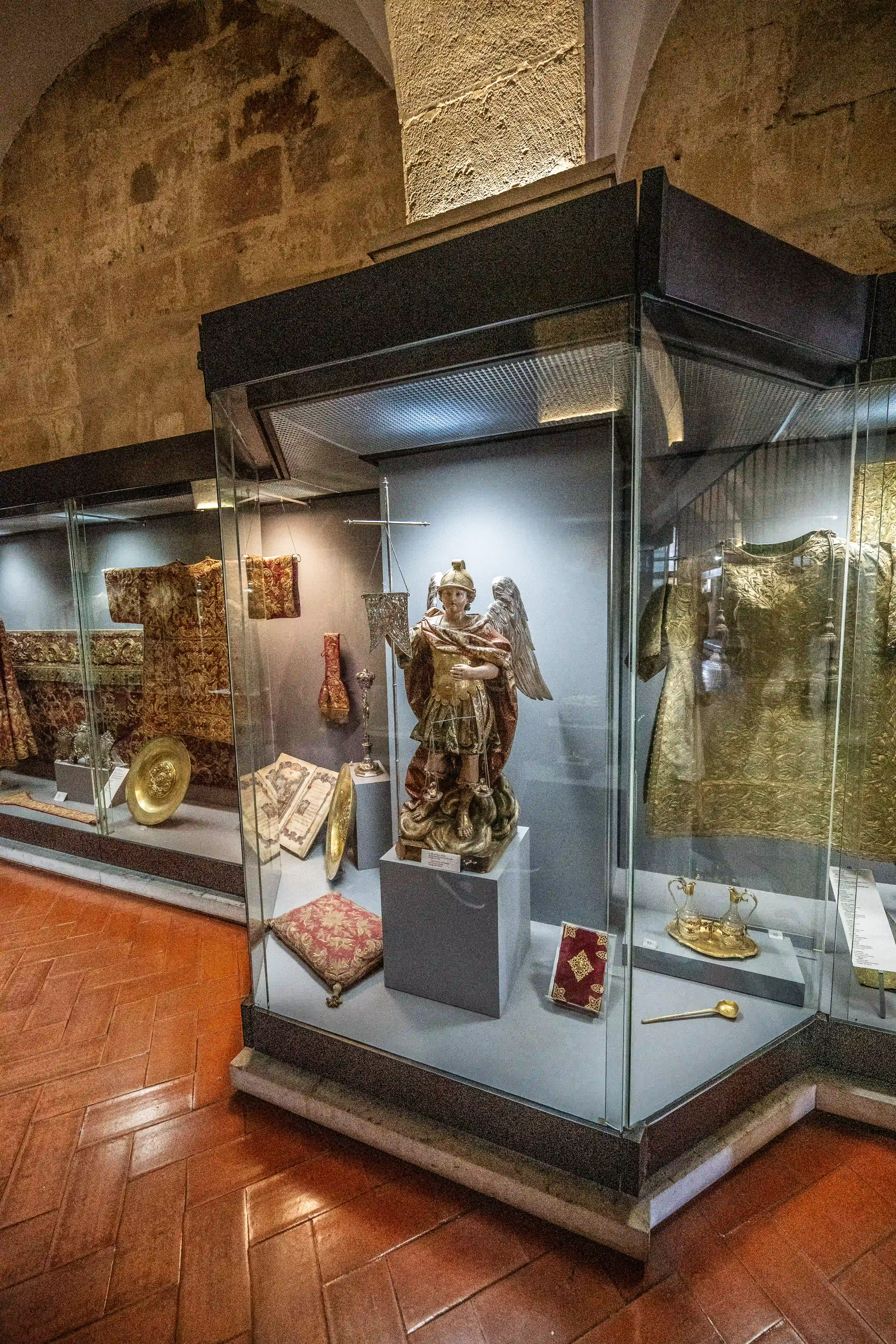 A museum display case containing a gilded statue of an angel with wings, elaborate religious garments, a golden dish, an ornate book, and other artefacts, set against a stone wall.
