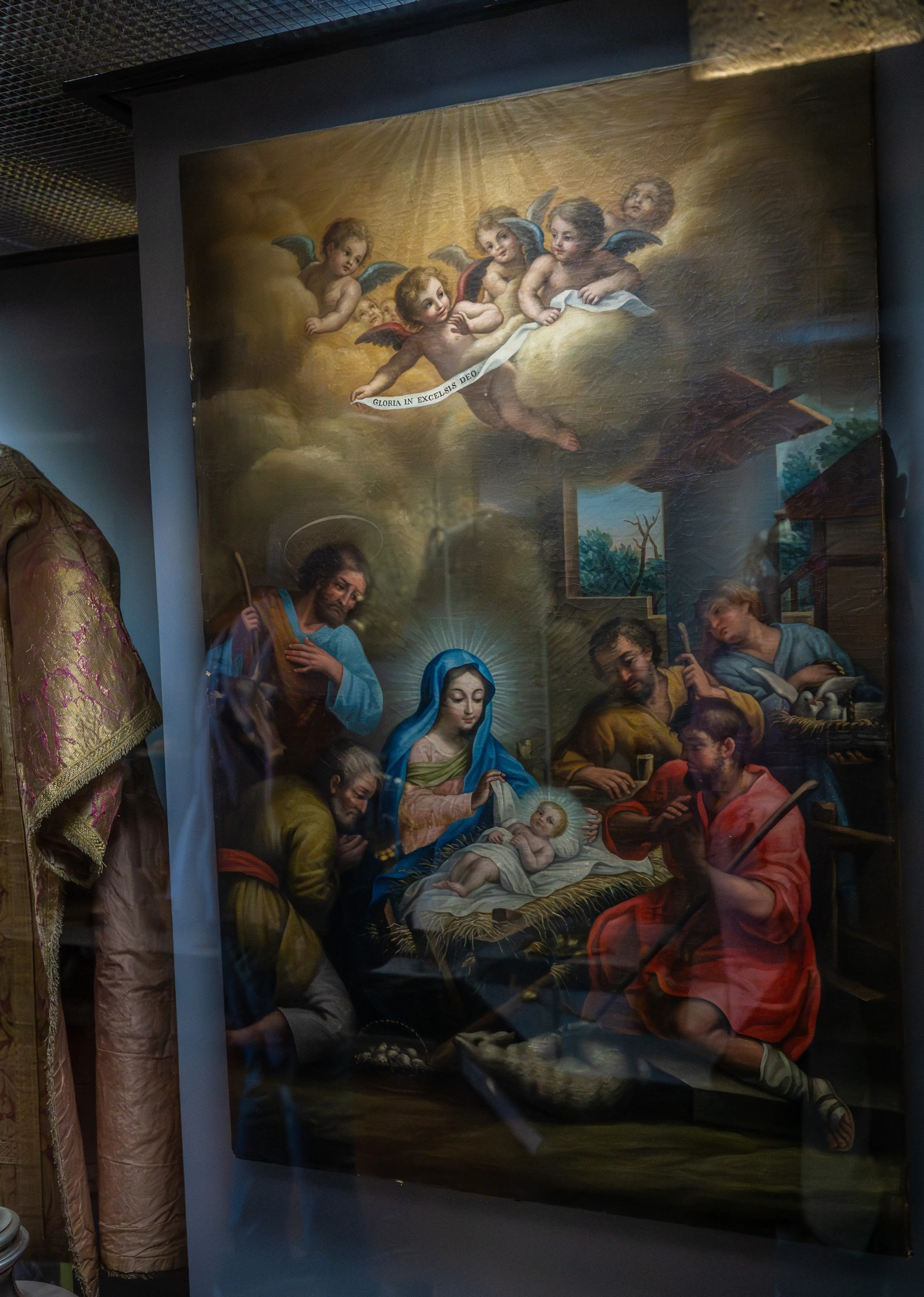 An oil painting of the Nativity scene, featuring the Virgin Mary, baby Jesus, Joseph, shepherds, and angels bearing a banner.
