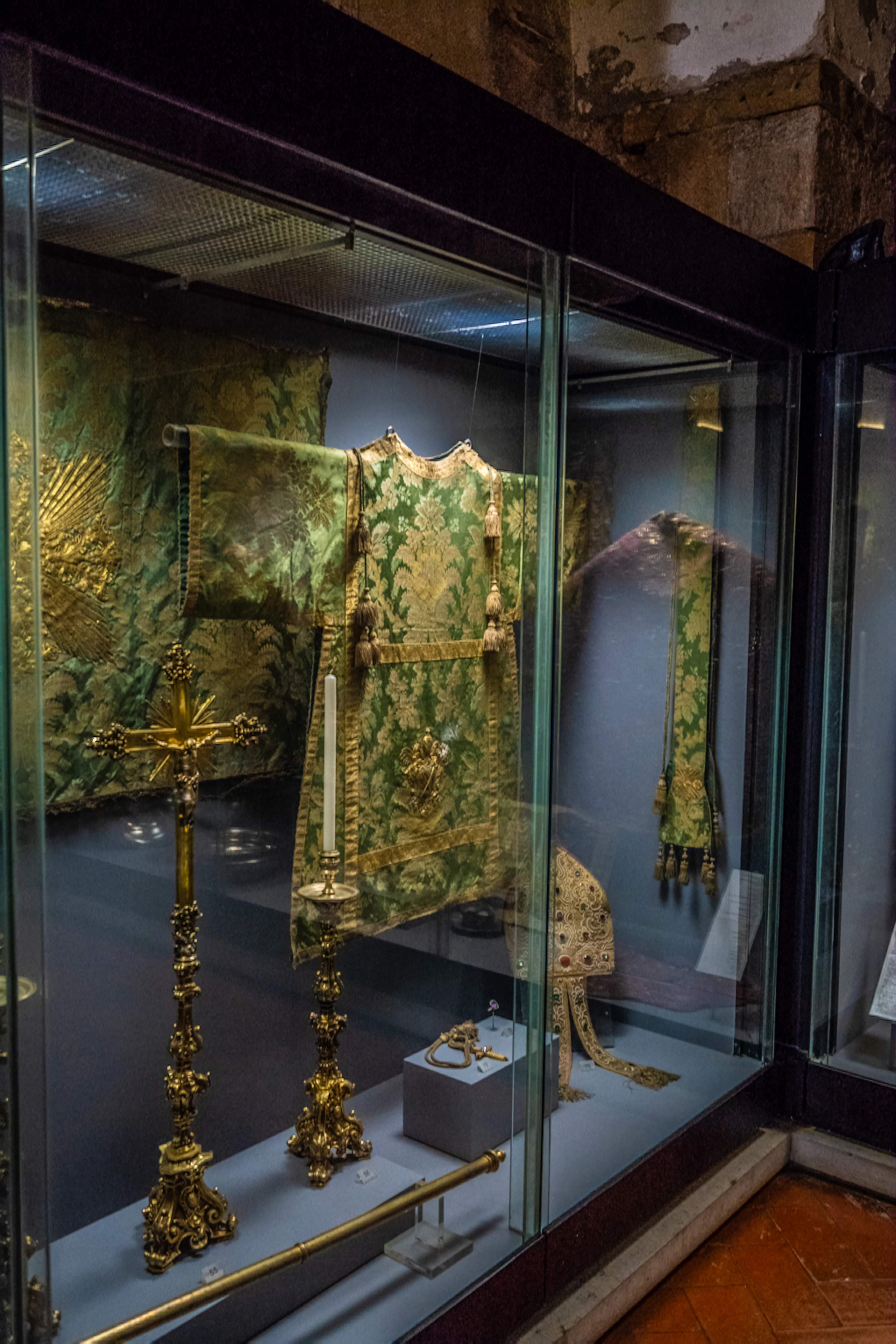 A museum display case featuring historical religious items, including a richly embroidered green chasuble, a golden crucifix, a candlestick, and a mitre.