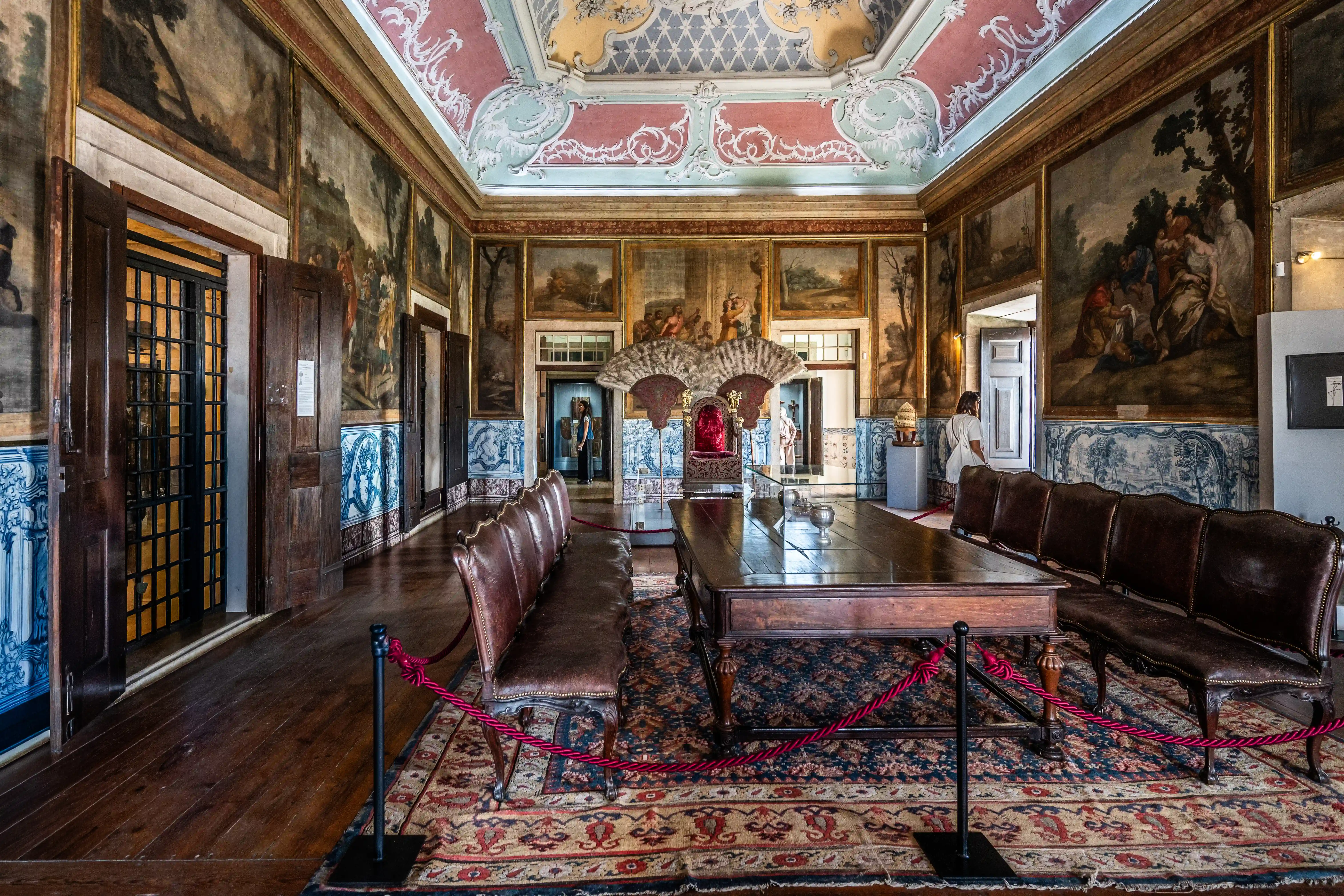 A wide shot of a richly decorated historic hall, showcasing a large wooden table, leather seating, a throne, and walls adorned with paintings and blue and white tiles.