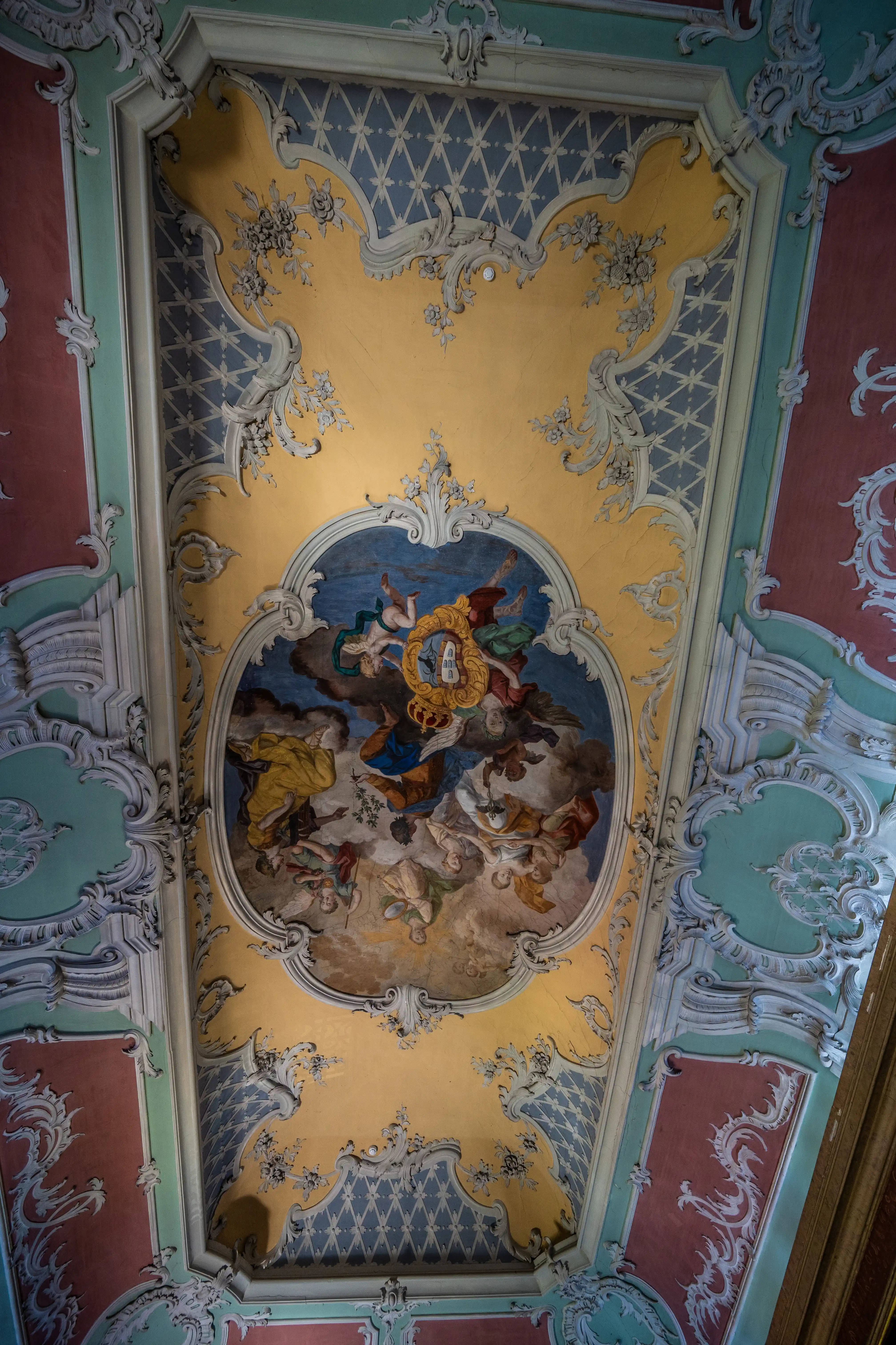 An ornate Baroque ceiling with a central fresco of figures in the sky, surrounded by intricate plasterwork in yellow, blue, rose, and teal.