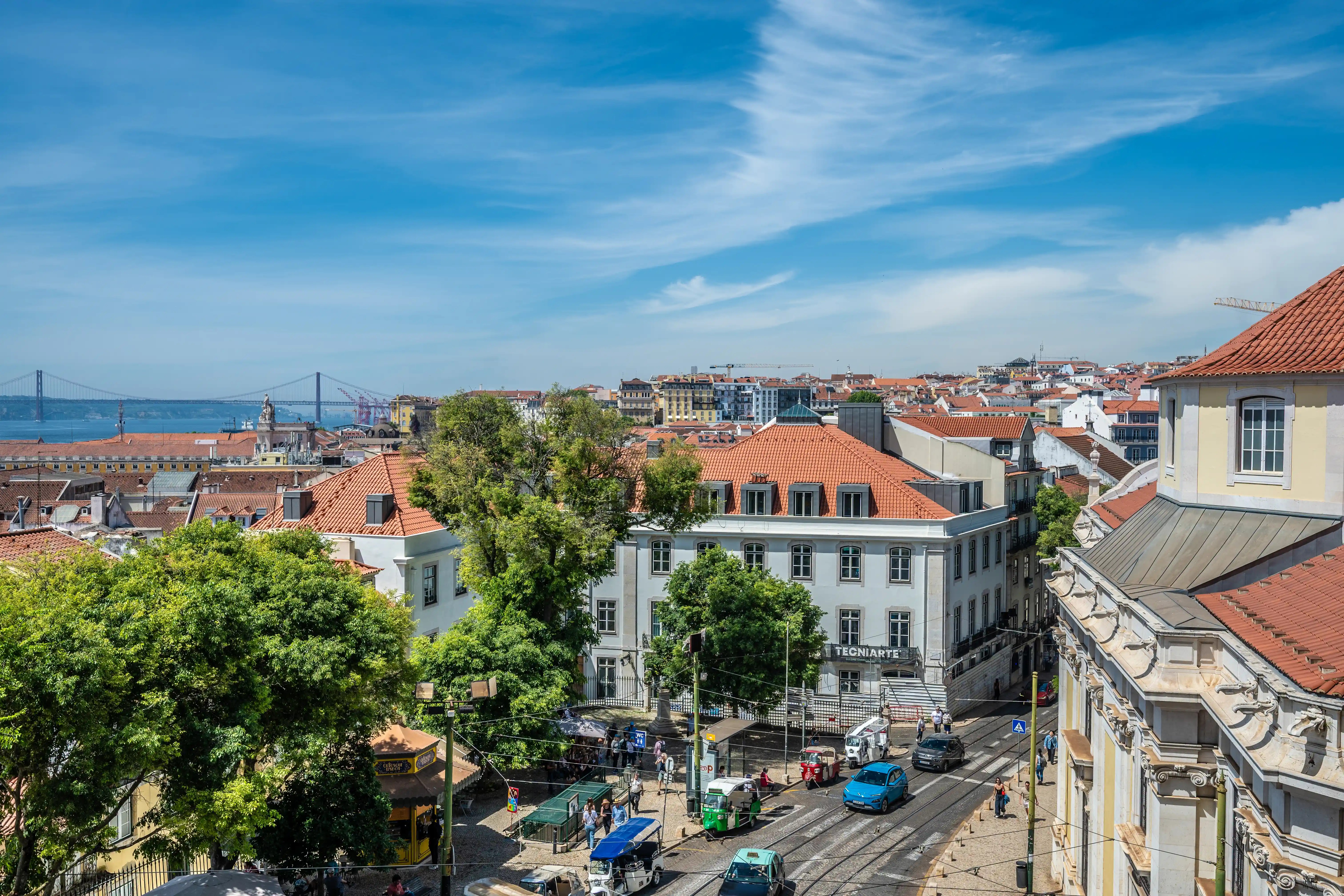 Aerial view of Lisbon's urban landscape with red-tiled roofs, a street scene with traffic, and the 25 de Abril Bridge visible across the river.
