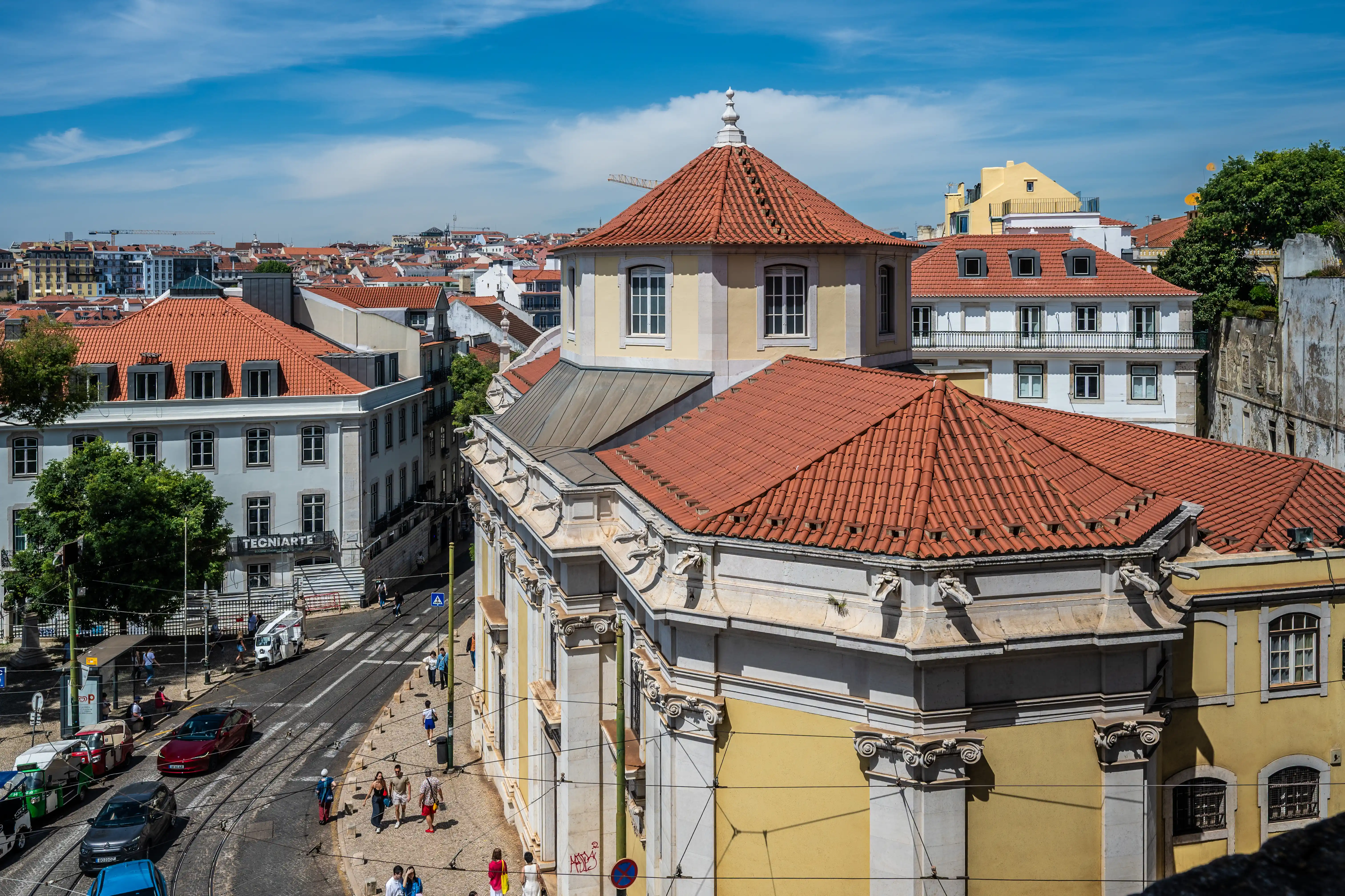  A high-angle shot of a street in Lisbon, Portugal, showing numerous terracotta-tiled roofs of old buildings. In the foreground, a yellow building with classical architectural elements stands out. Cars, trams, and pedestrians are visible on the street below. The sky is blue with white clouds.