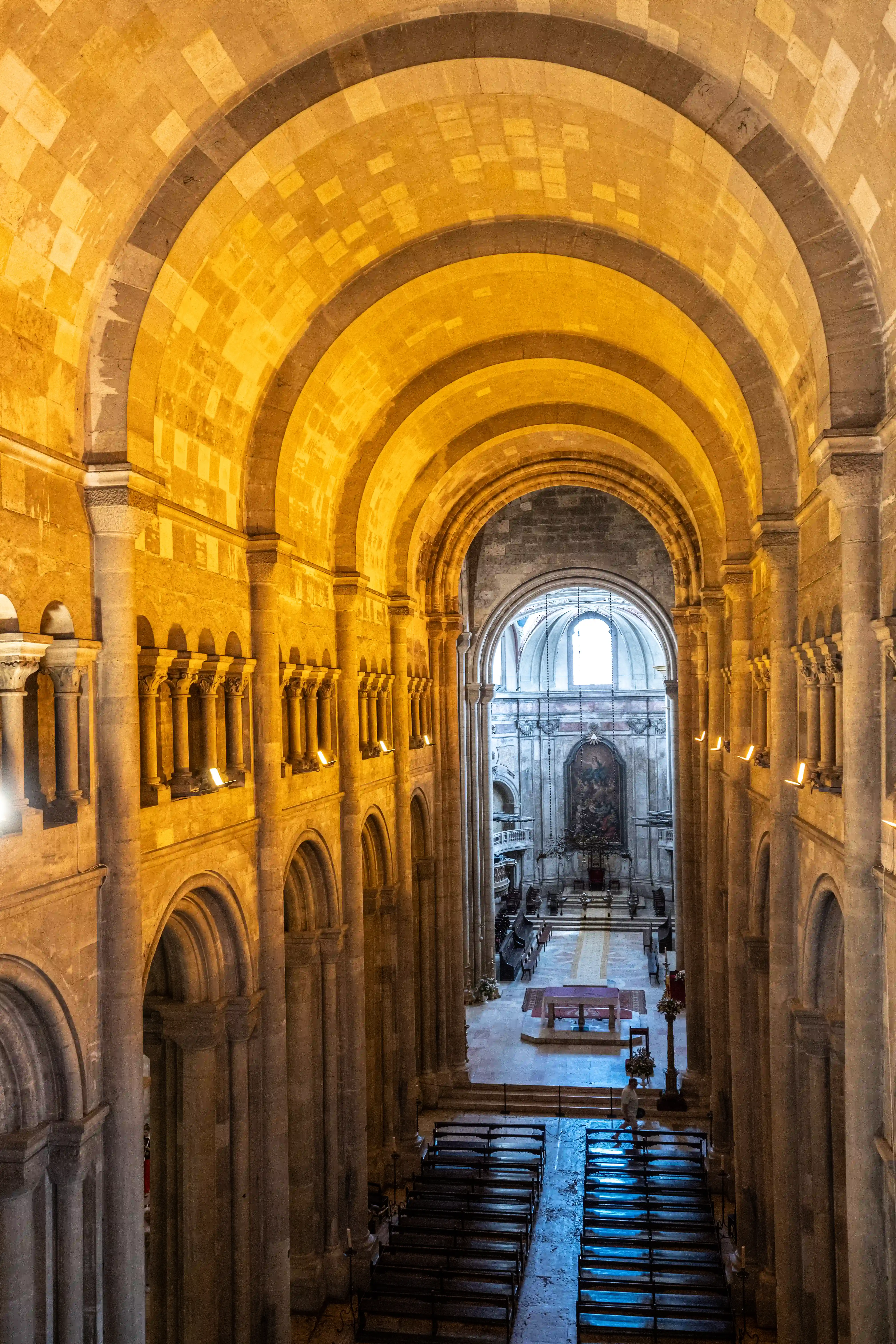  Interior view of a large stone cathedral with multiple rounded arches receding into the distance, a lit altar at the far end, and rows of pews.