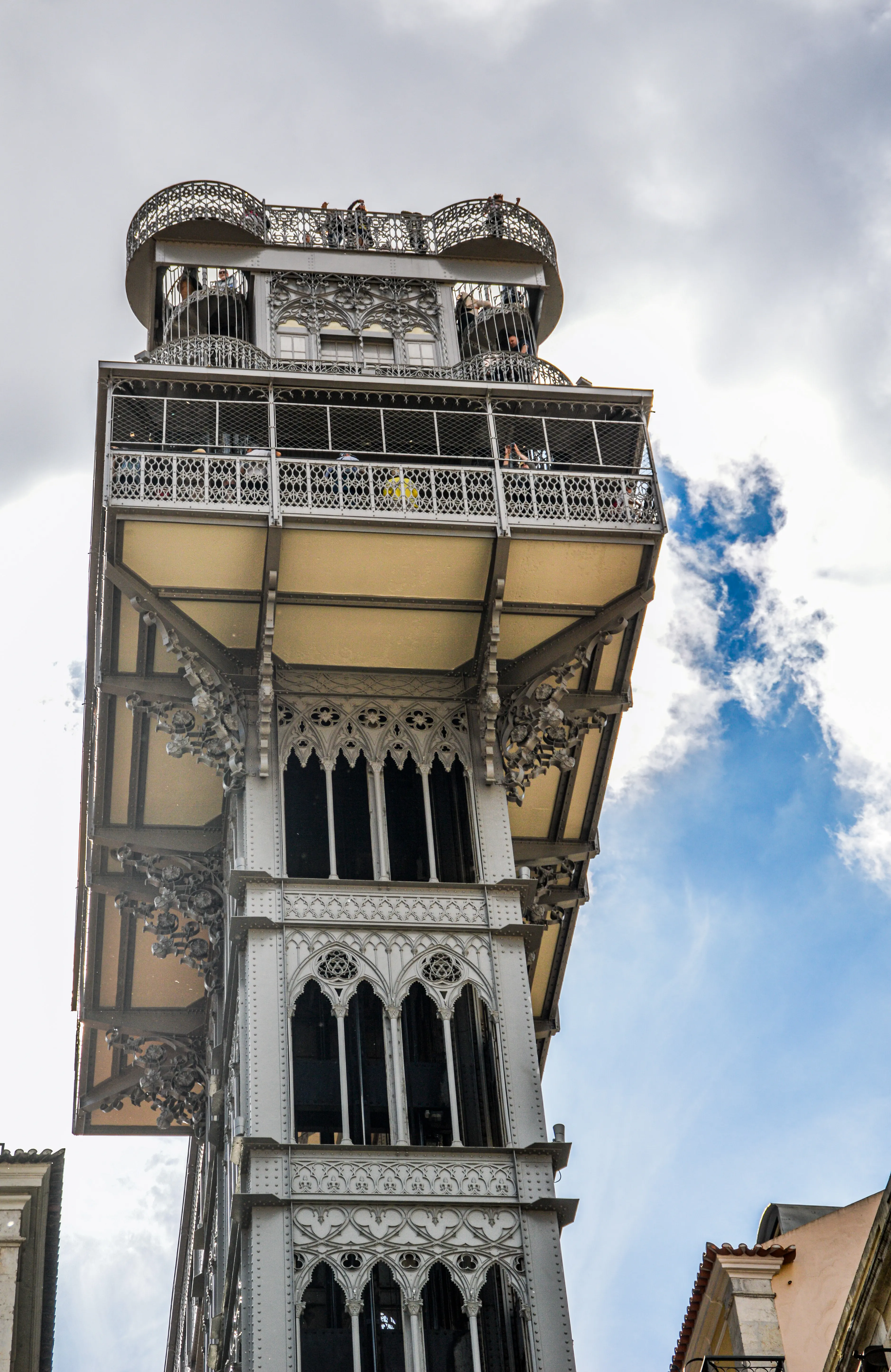 Close-up, low-angle view of the historic Santa Justa Lift in Lisbon, Portugal, showcasing its detailed ironwork, gothic windows, and observation decks filled with people, under a dynamic cloudy sky.