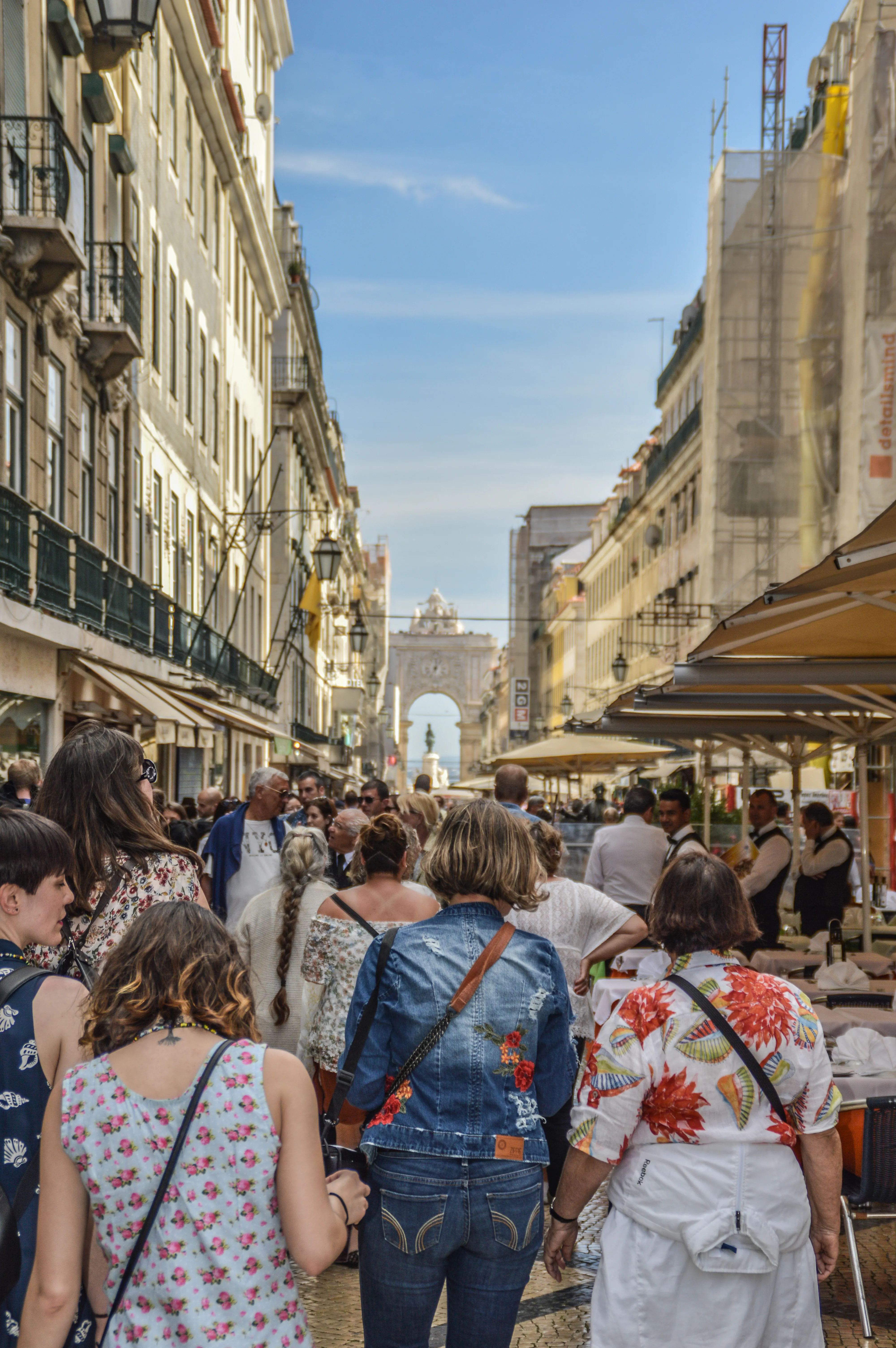 A crowd of people walk down a sunlit street in Lisbon, Portugal, with tall buildings on either side. A large ornate archway stands at the end of the street under a blue sky.