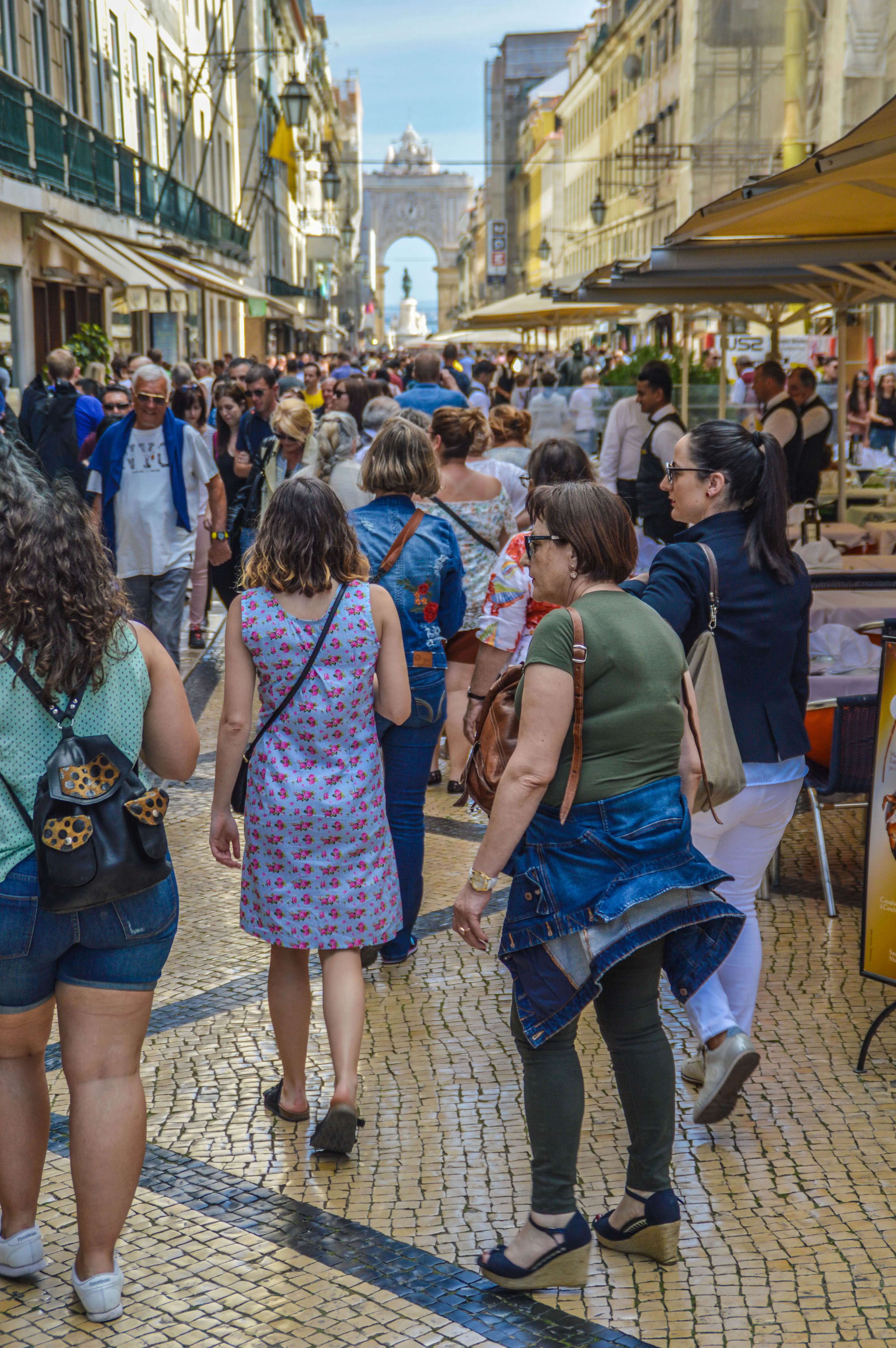 A lively street scene with many people walking on a patterned cobblestone path. Buildings with balconies and awnings line both sides of the street. In the background, a large ornate archway stands under a blue sky.