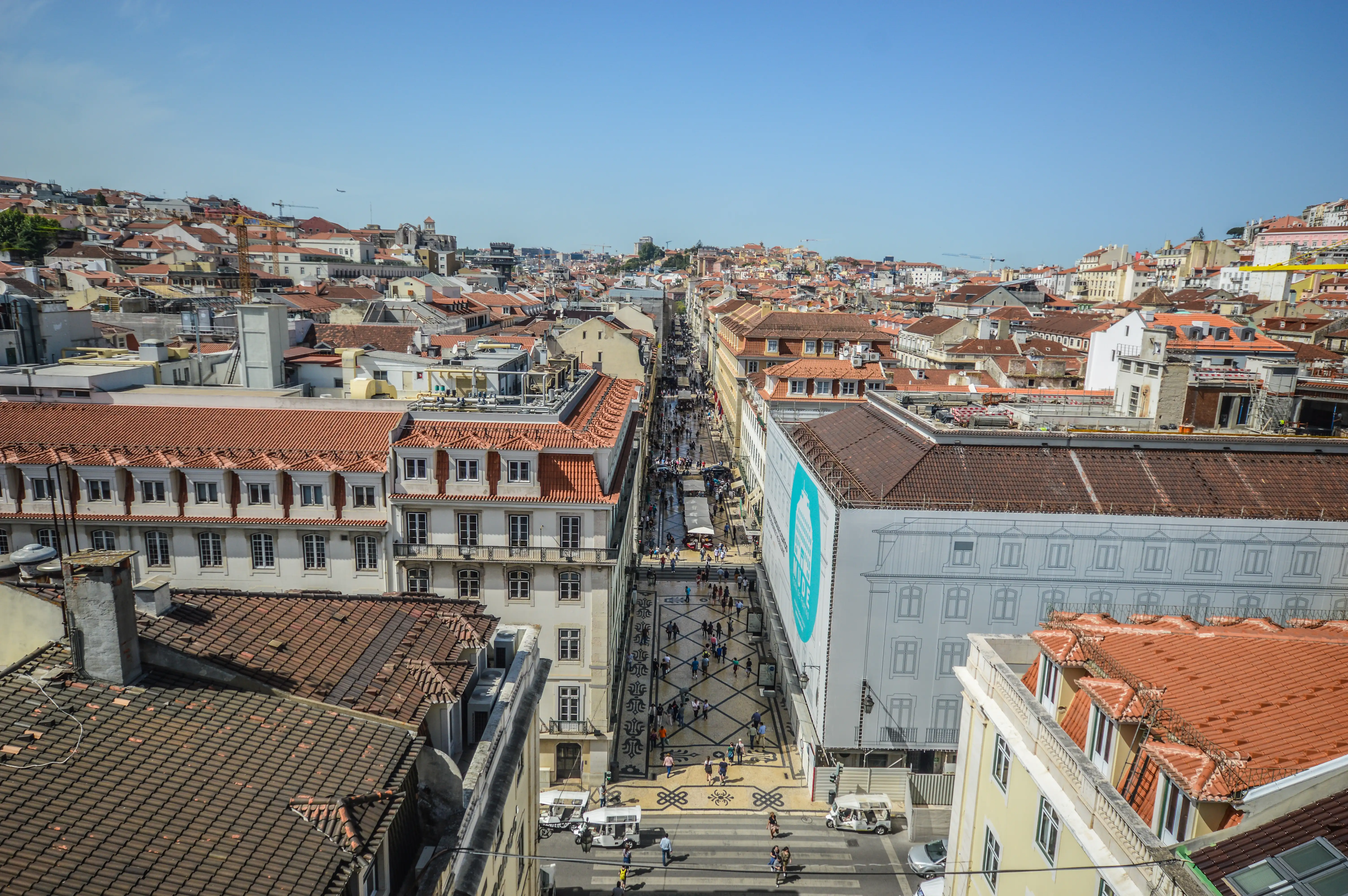  A high-angle shot shows a busy street in Lisbon, Portugal, with buildings on either side covered in red-tiled roofs. Pedestrians fill the street, and a mosaic pattern runs down the centre. White electric cars are visible near a zebra crossing. The sky is clear and blue.
