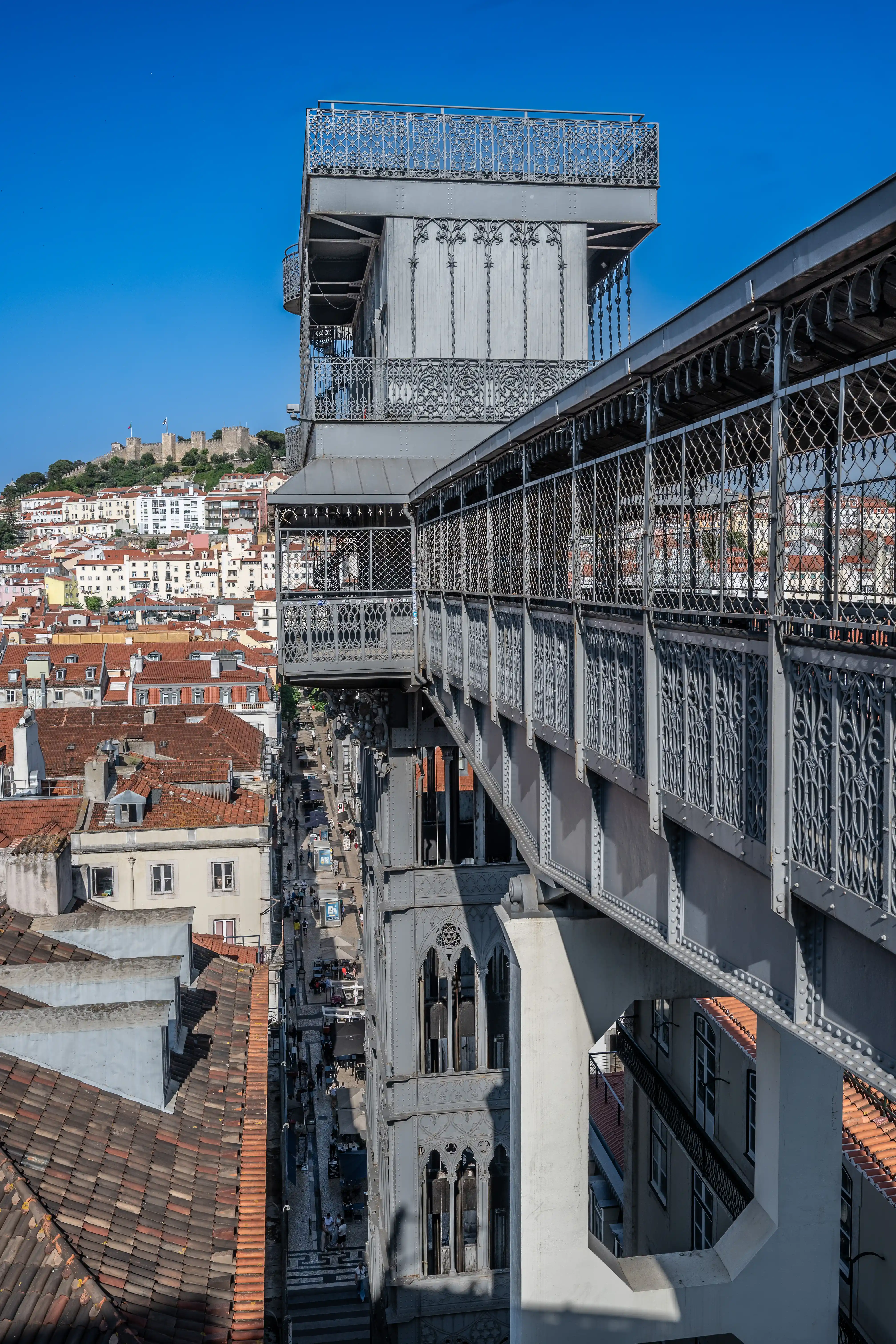 The Santa Justa Lift, a tall, grey wrought-iron elevator structure, rises above the cityscape of Lisbon, Portugal. The lift features ornate metal railings and decorative patterns. In the distance, the city is filled with buildings with red-tiled roofs, and the ancient São Jorge Castle sits atop a hill under a bright blue sky.