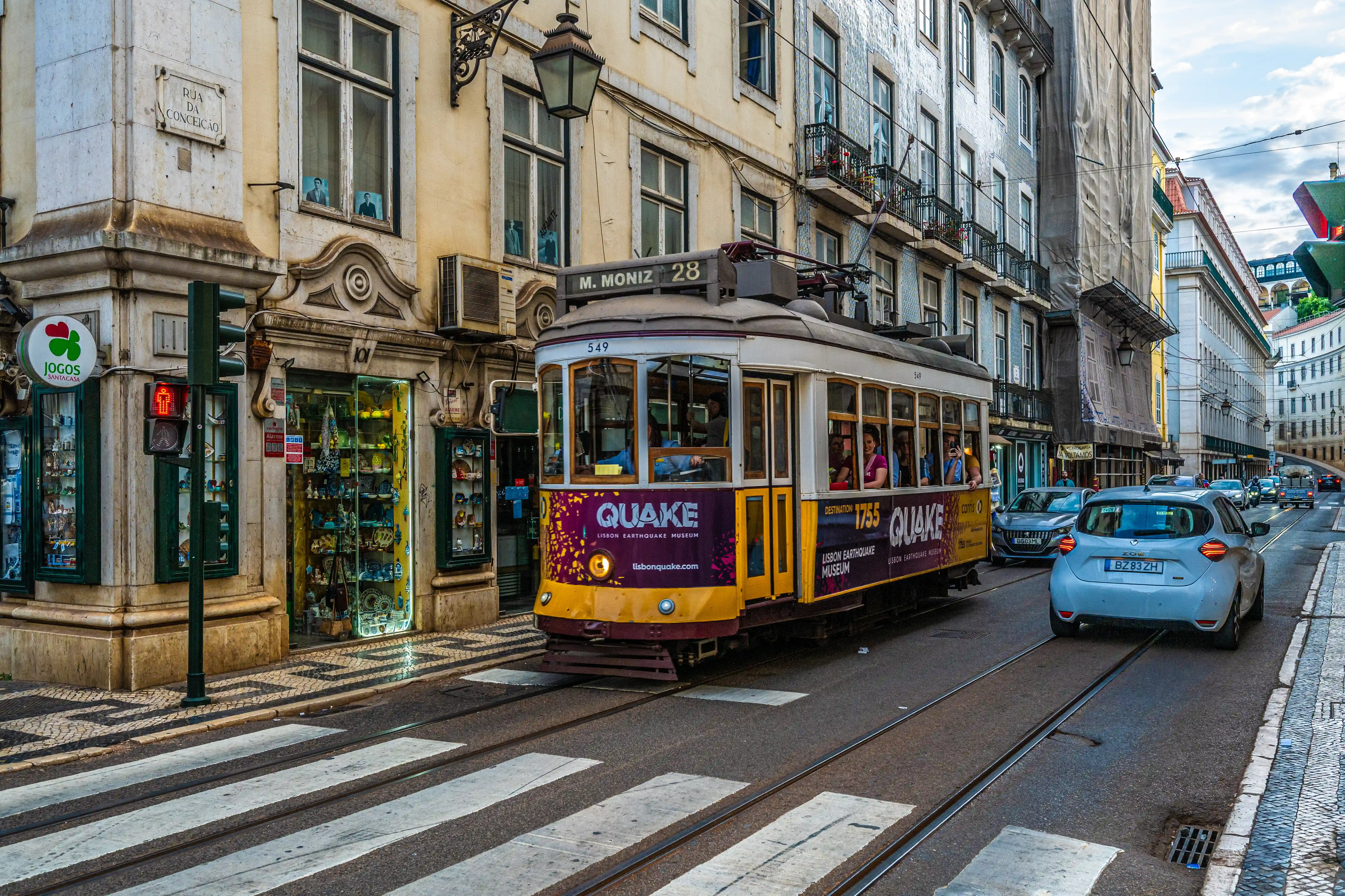 A yellow Lisbon tram with the number 549 and an advertisement for the Lisbon Earthquake Museum (Quake) travels along tram tracks on a street in Lisbon. Pedestrians are visible inside the tram, and a white car follows behind. Historic buildings with shops line the street.