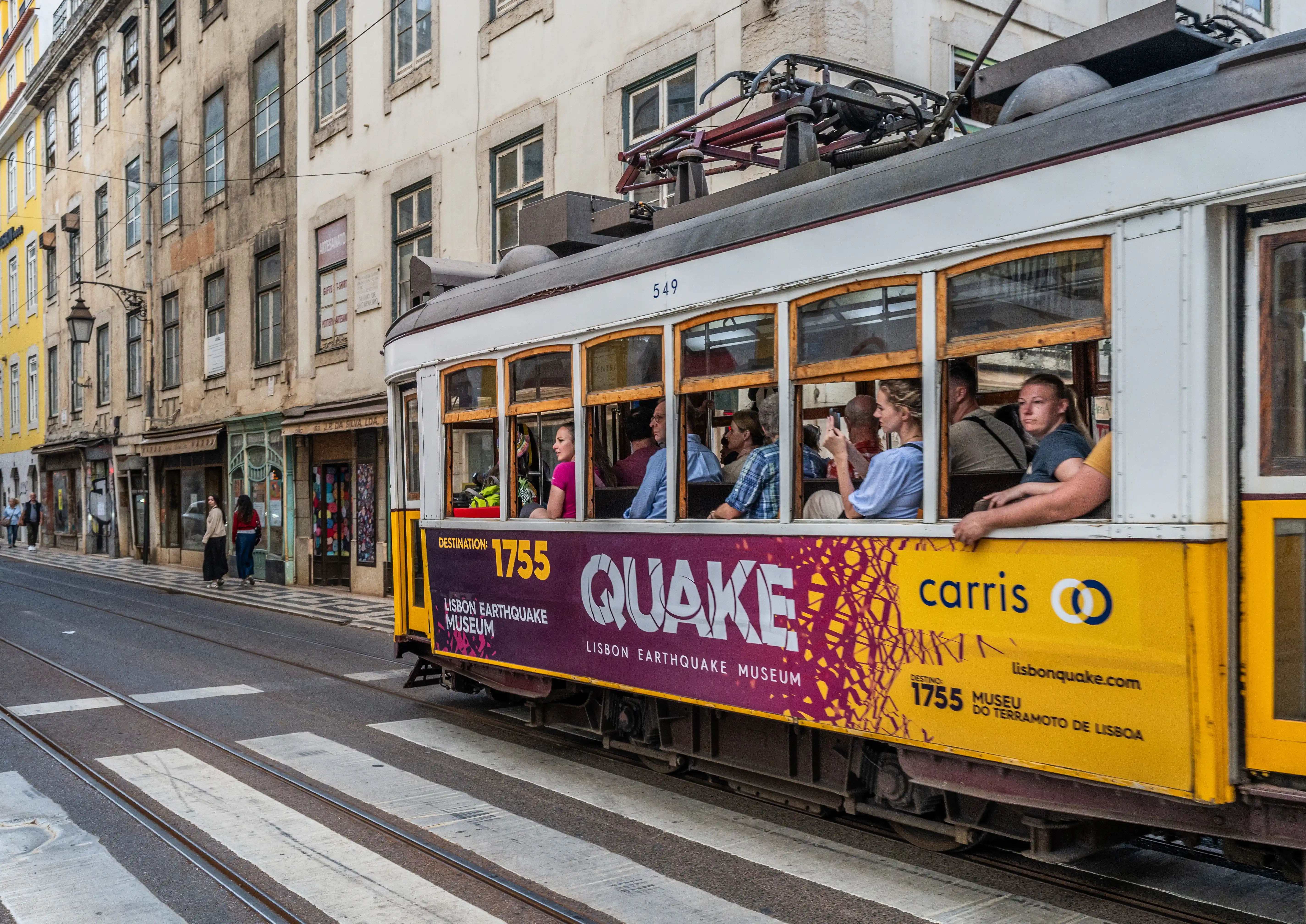 A yellow tram with passengers inside travels past old buildings, featuring an advertisement for the 