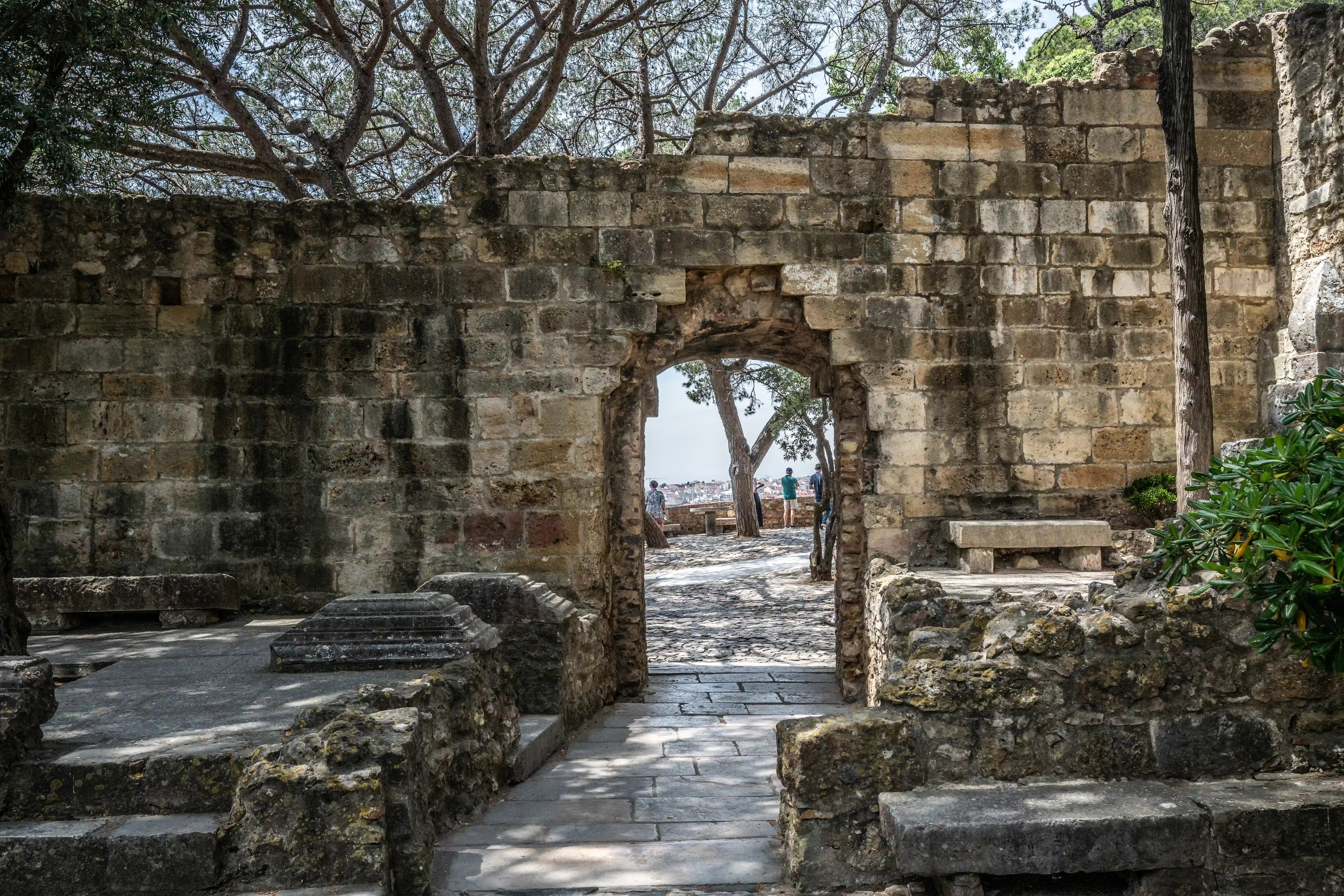 An arched opening in an old stone wall, revealing a sunlit path with scattered stone structures and people in the distance, framed by trees.