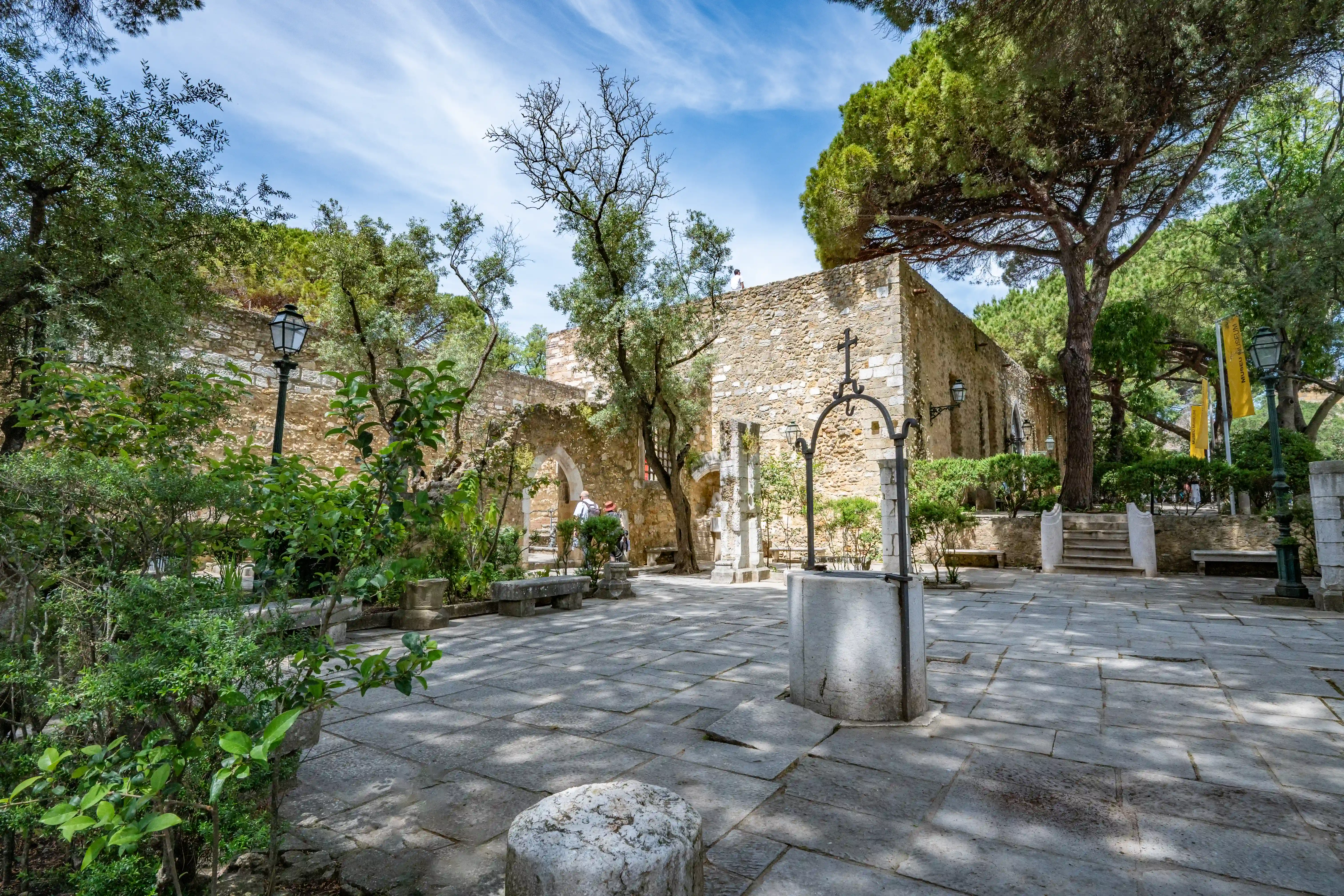 A serene view of a historic courtyard featuring an old stone well with a wrought-iron cross, surrounded by ancient stone walls, trees, and paved ground. The scene evokes a sense of timelessness and historical depth.