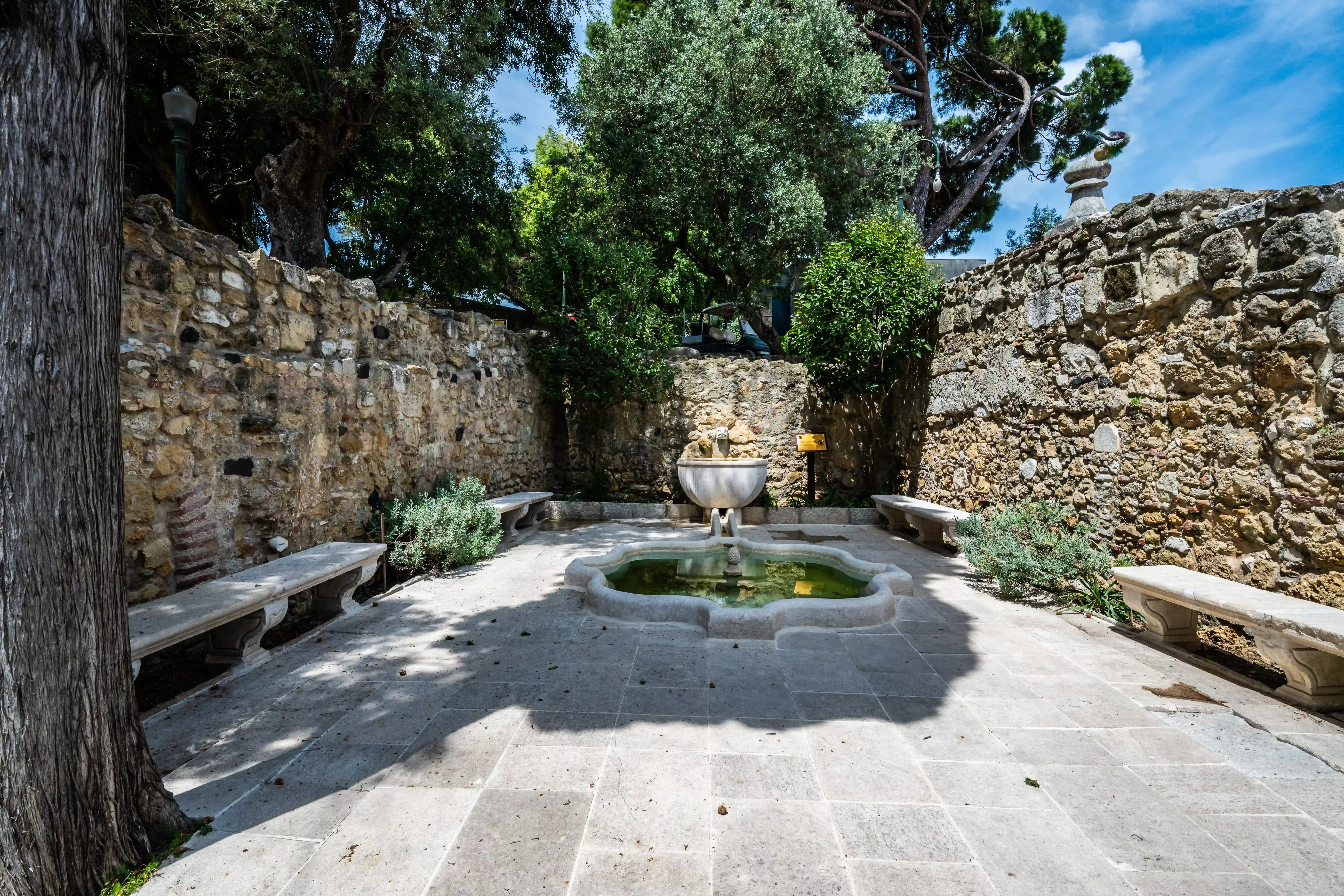 A courtyard scene with a stone fountain, stone benches, and stone walls, surrounded by trees.