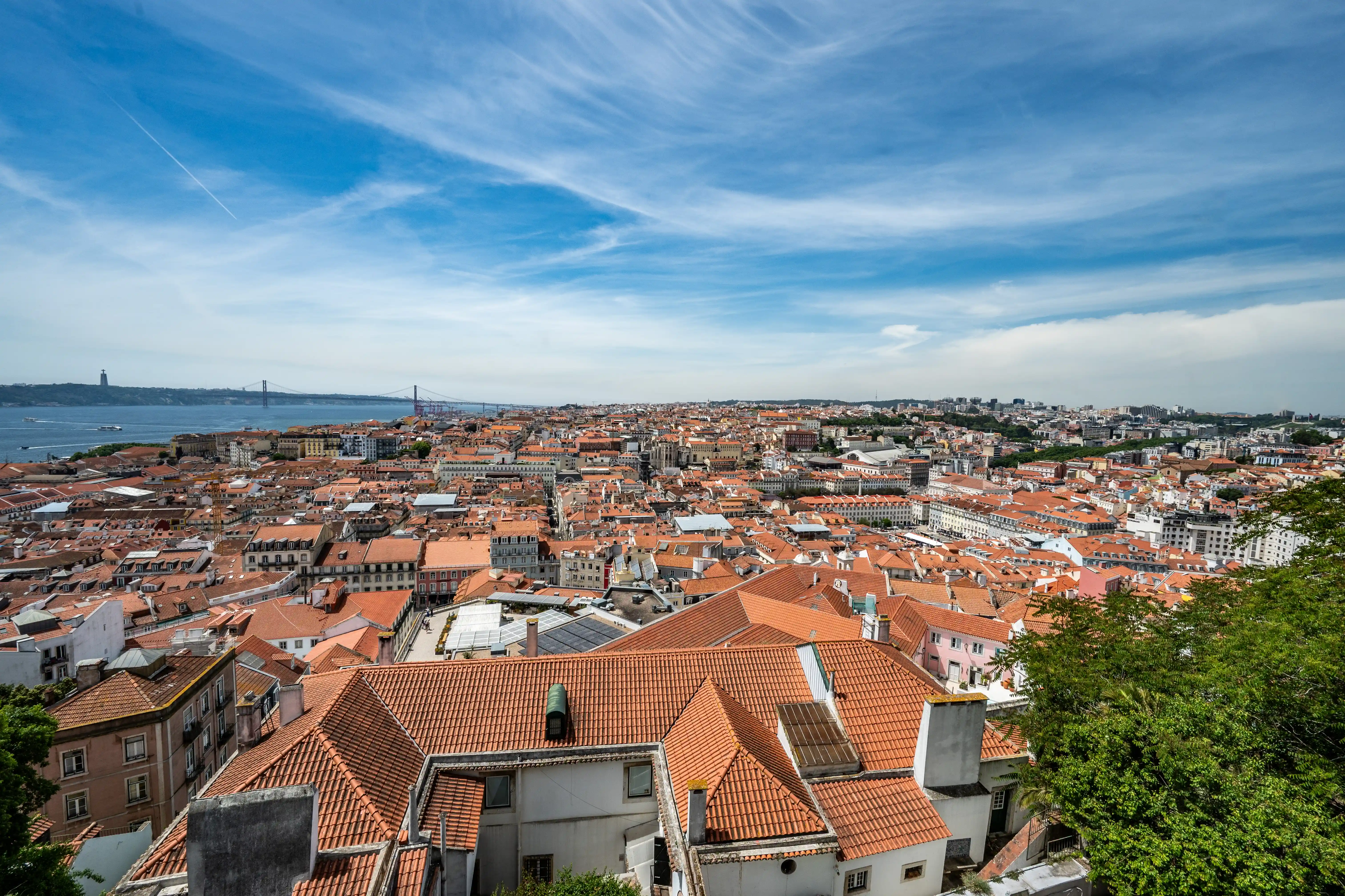 An aerial view of Lisbon, Portugal, showcasing a vast expanse of red-tiled roofs under a blue sky. A suspension bridge crosses the Tagus River in the distance, with a monument visible on the opposite bank.