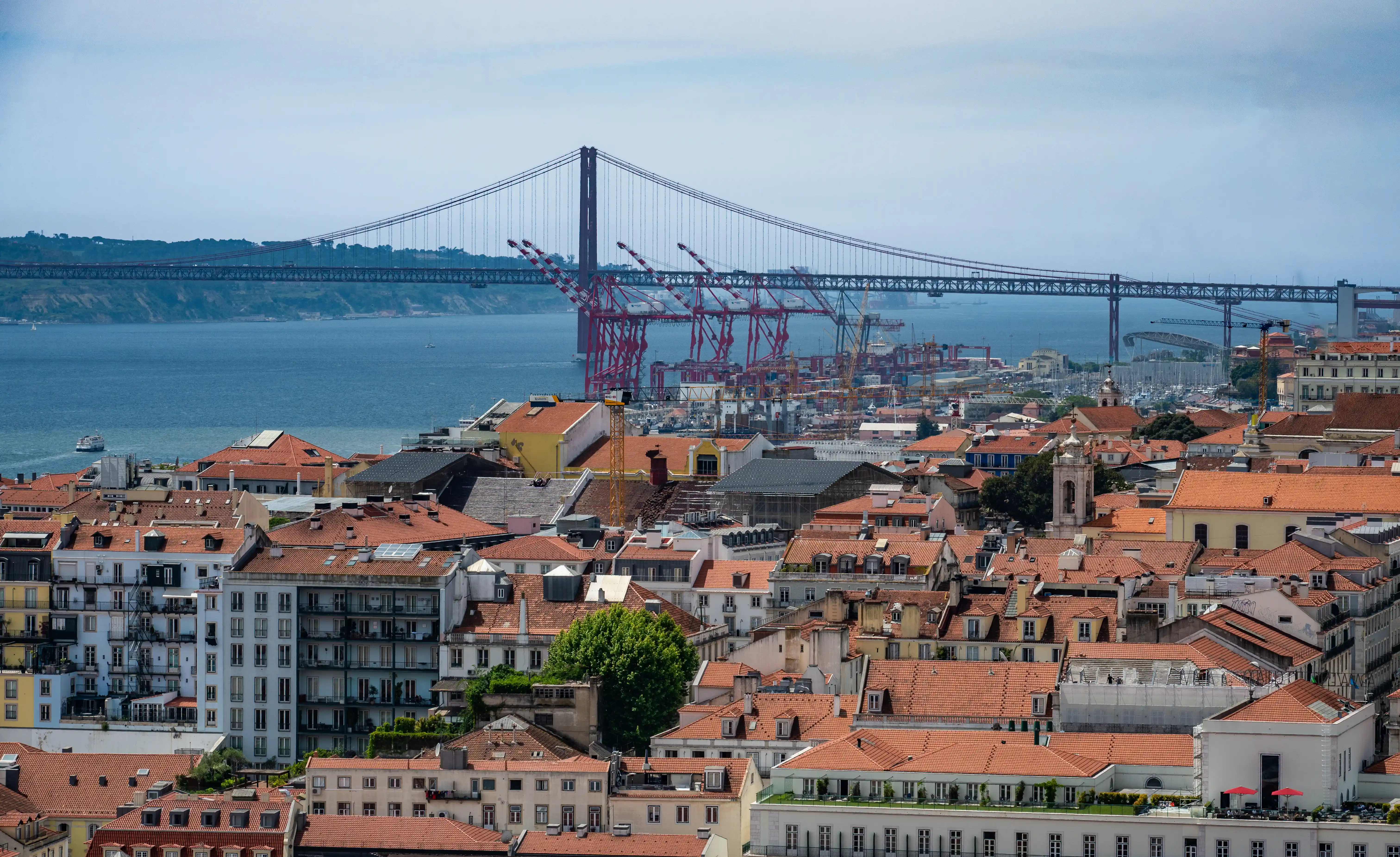 Wide shot of Lisbon, Portugal, showing the 25 de Abril Bridge and port in the distance, with a dense arrangement of red-tiled rooftops in the foreground.