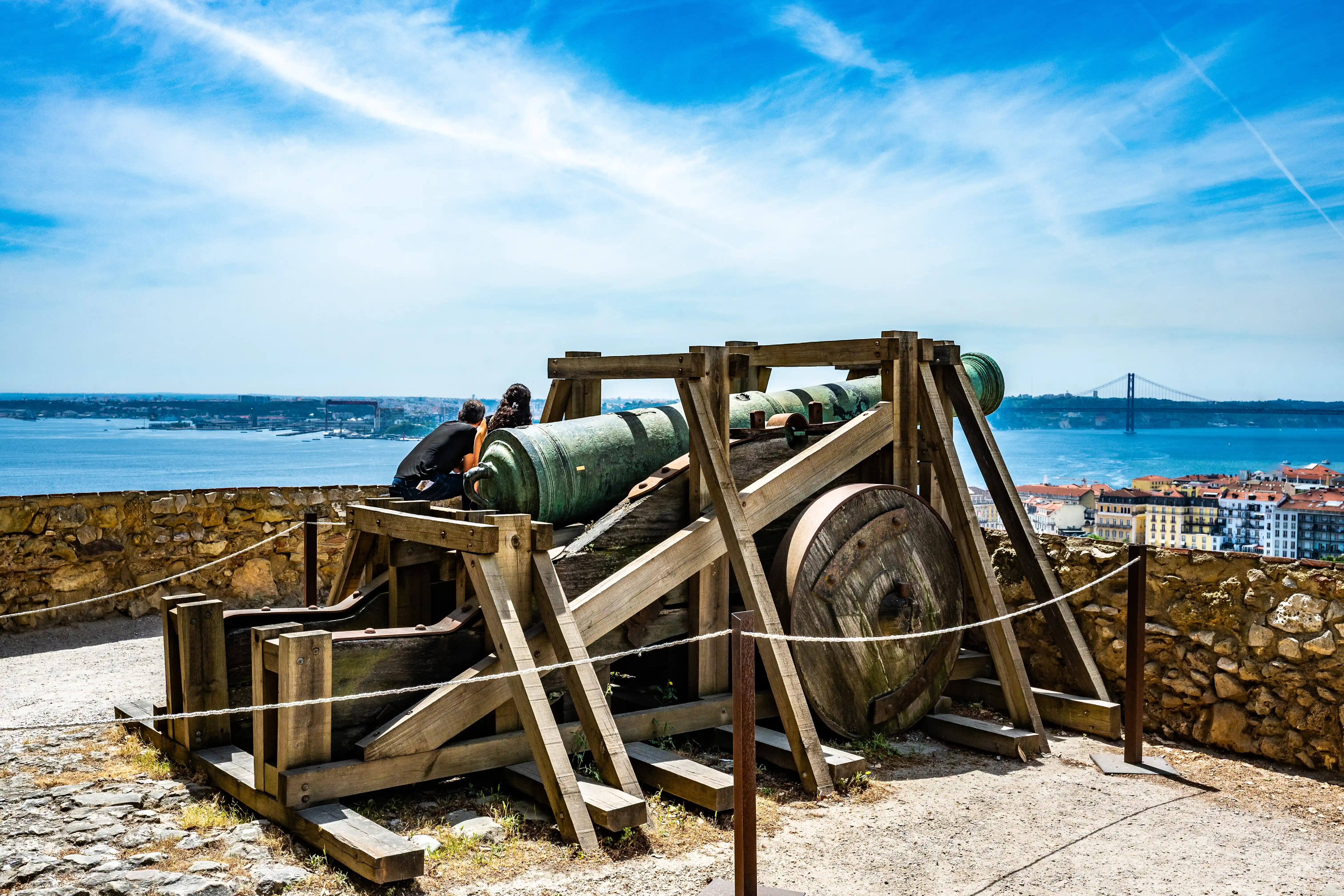 Two people observing a large, old cannon on a wooden mount, with a river, city, and bridge in the background under a bright sky.
