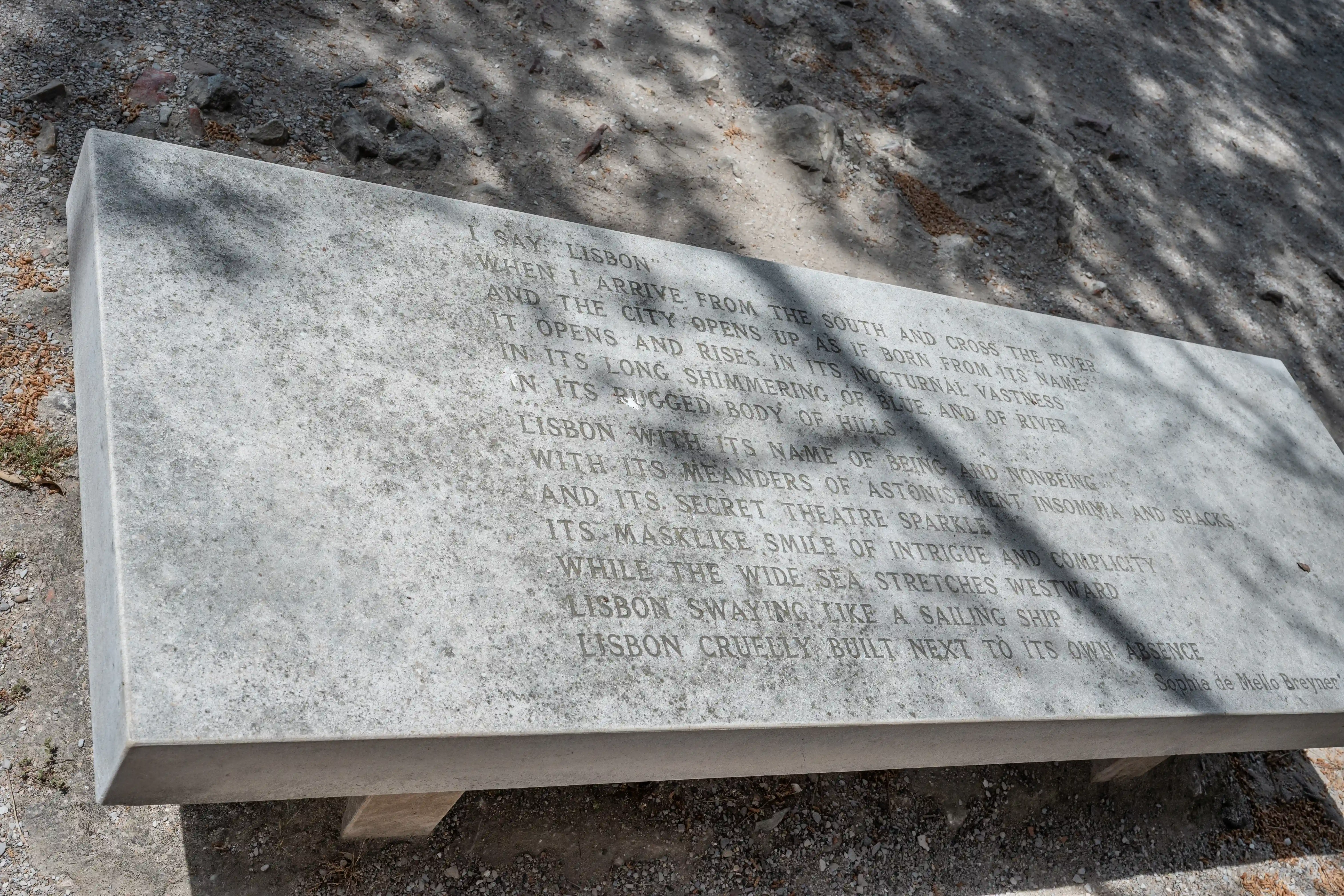 A light grey stone bench carved with a poem, detailing the city of Lisbon, set outdoors on a gravel path under dappled sunlight.