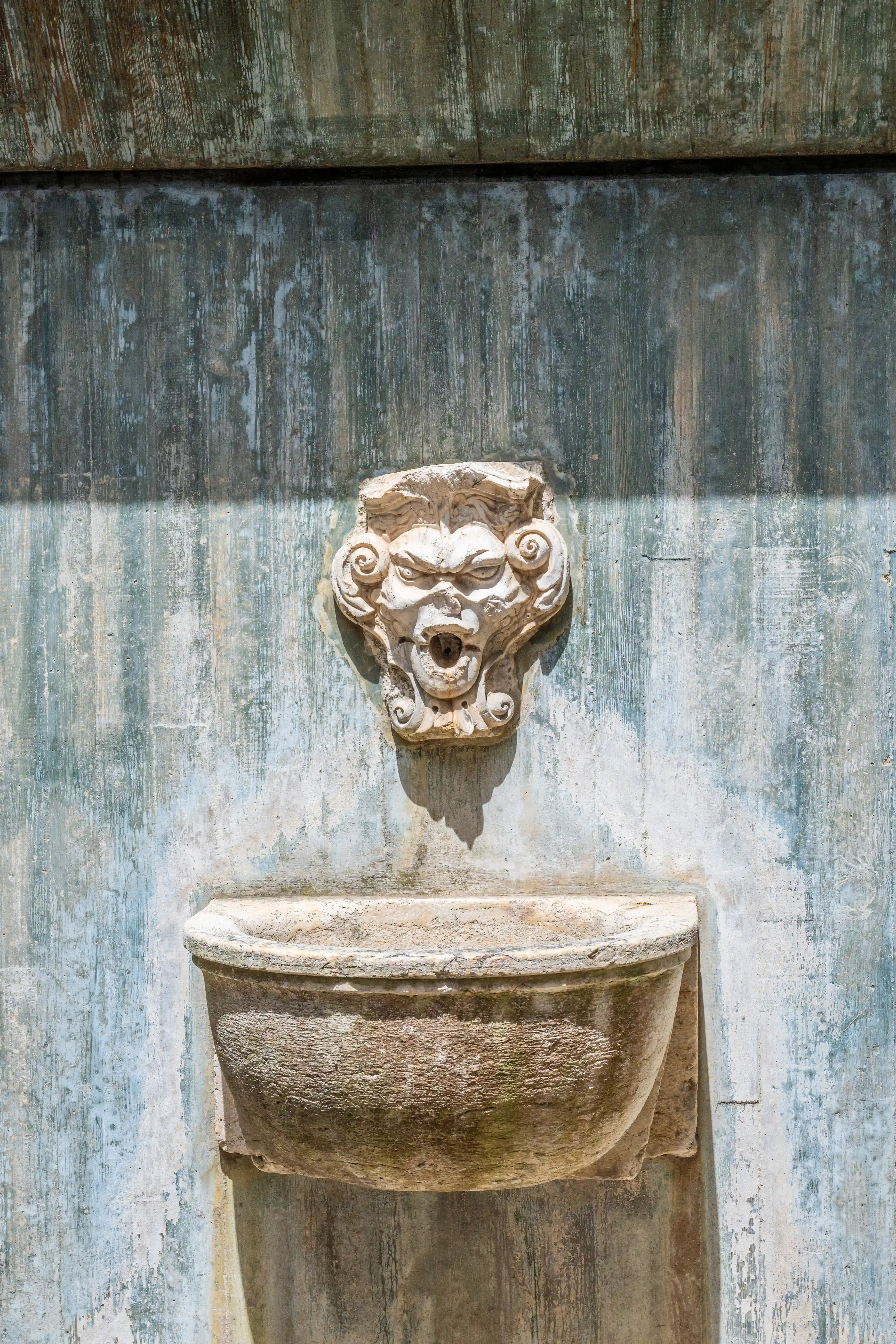A weathered stone gargoyle fountain with an open mouth and a basin below, mounted on a textured, distressed wall with shadows cast across it.