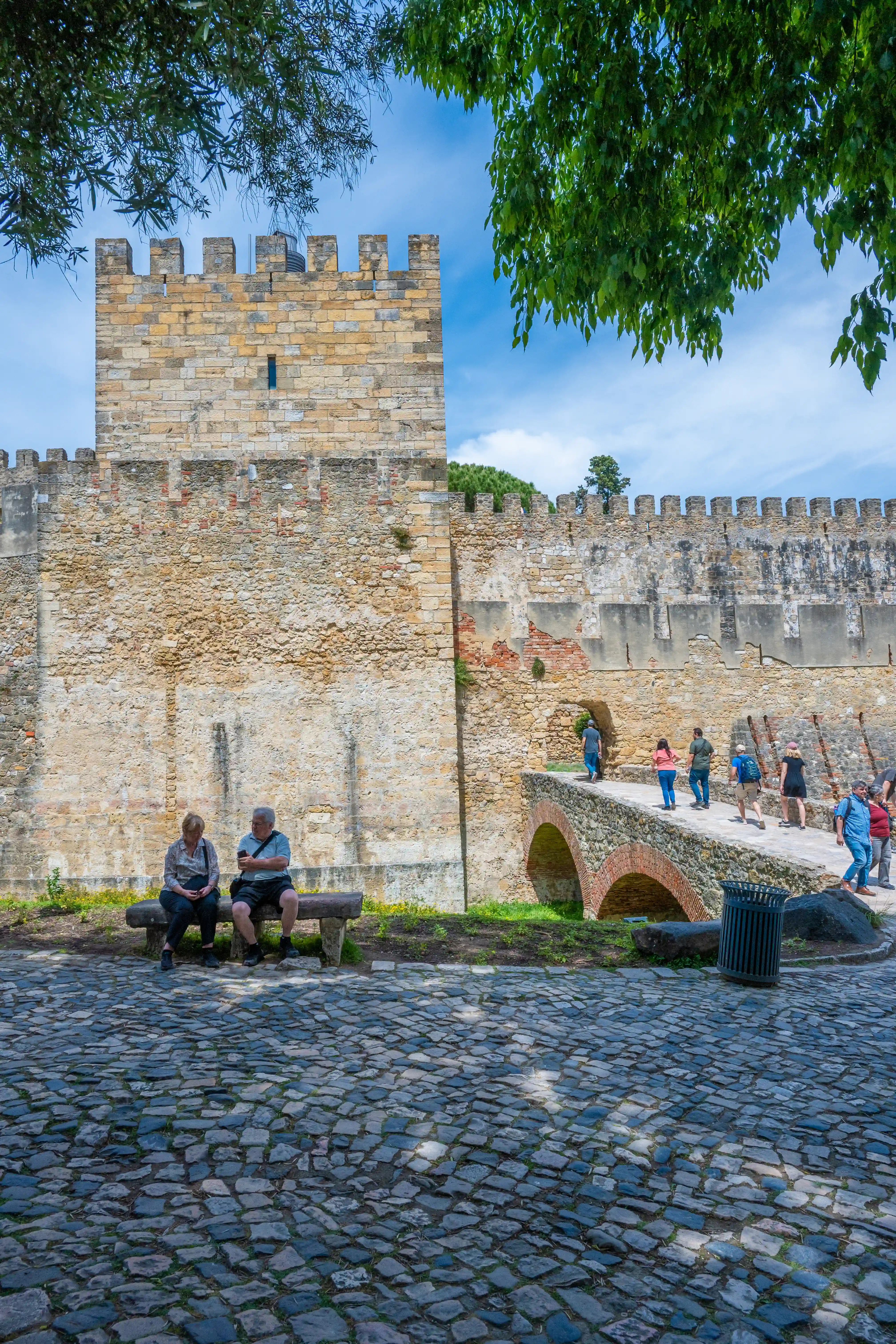 A stone castle wall with battlements under a cloudy sky, a cobblestone path in the foreground, two people sitting on a bench, and a group walking over a bridge.
