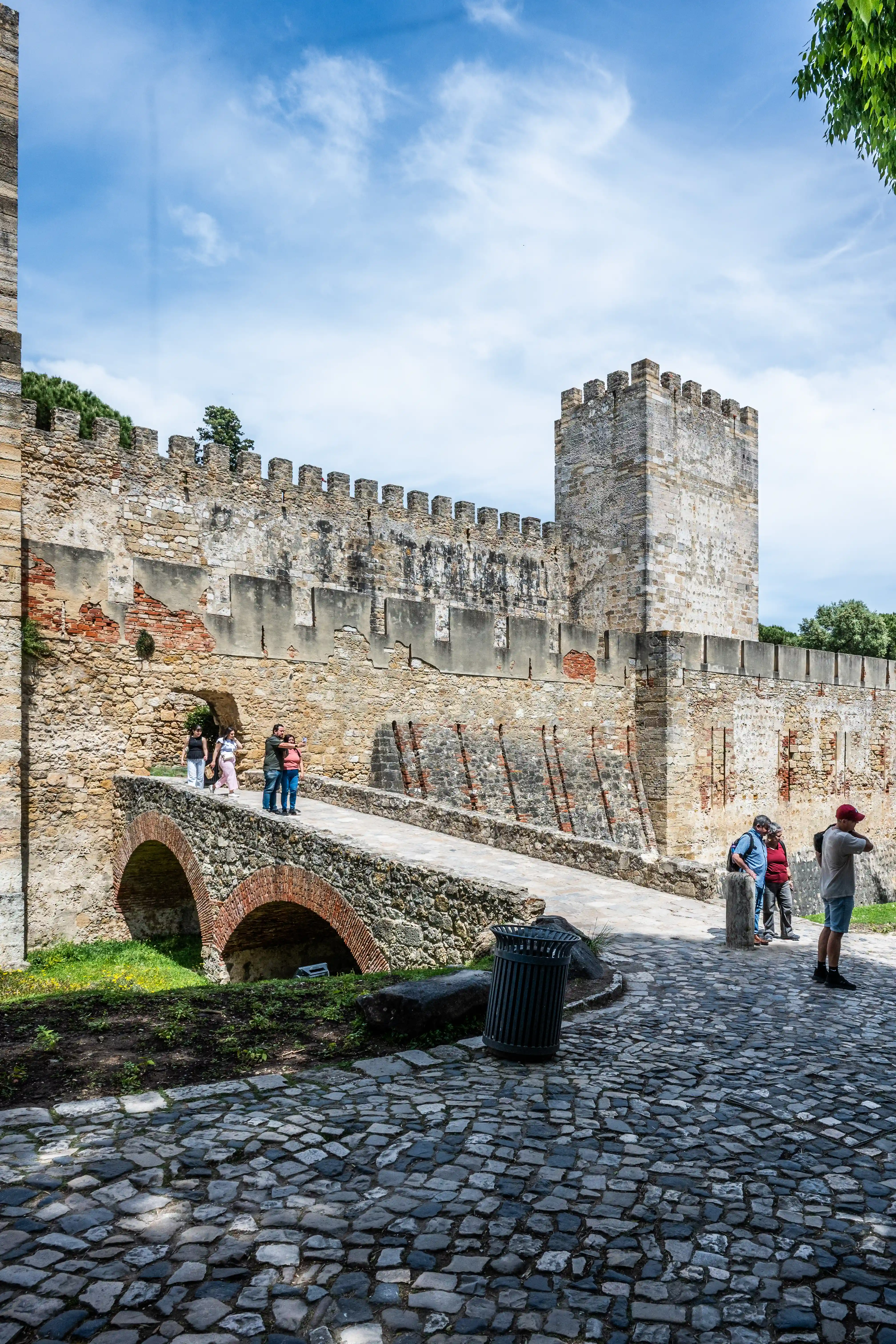 A stone bridge with arched foundations leads towards the weathered walls and a tower of an old castle. Several tourists are seen walking on the bridge and gathered near the entrance, whilst others stand on the cobbled courtyard. The sky above is partly cloudy, with patches of blue visible.