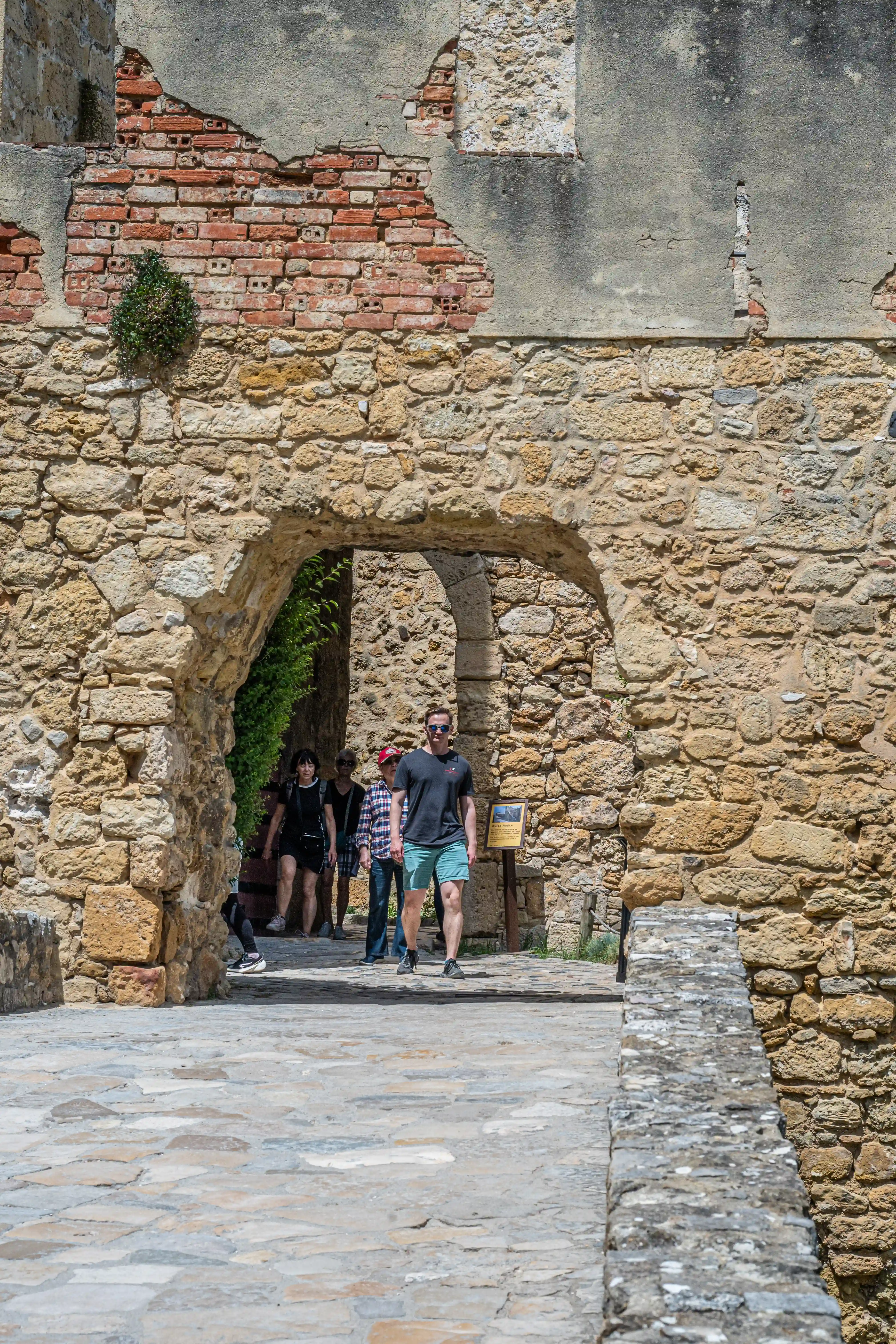 A man in a black t-shirt and blue shorts walks towards the camera through a stone archway, with other tourists behind him. The surrounding walls are made of rough stone with areas of crumbling plaster and brick.