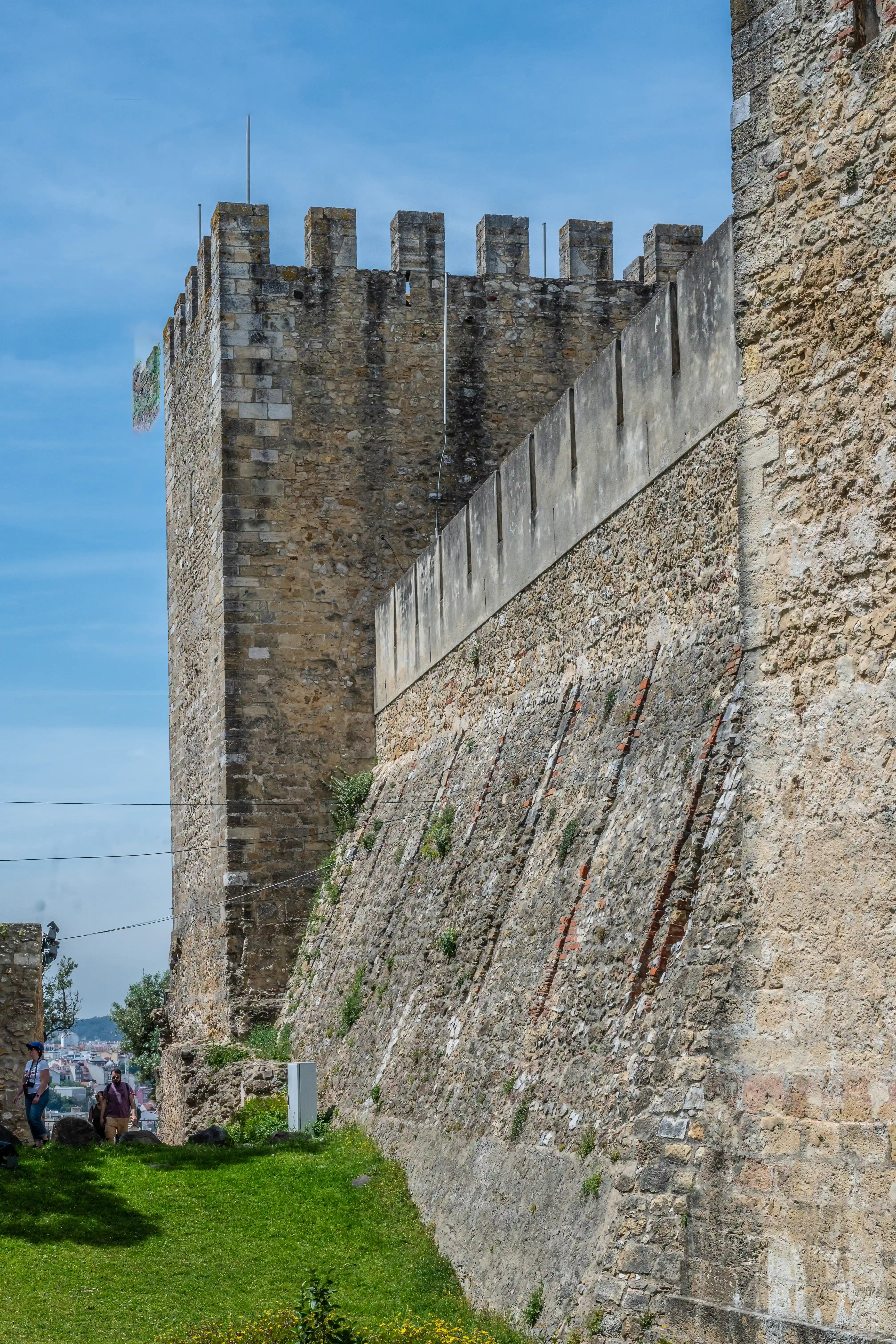 A tall stone tower and crenellated wall of an ancient fortress under a blue sky, with a green lawn and distant city in the background.