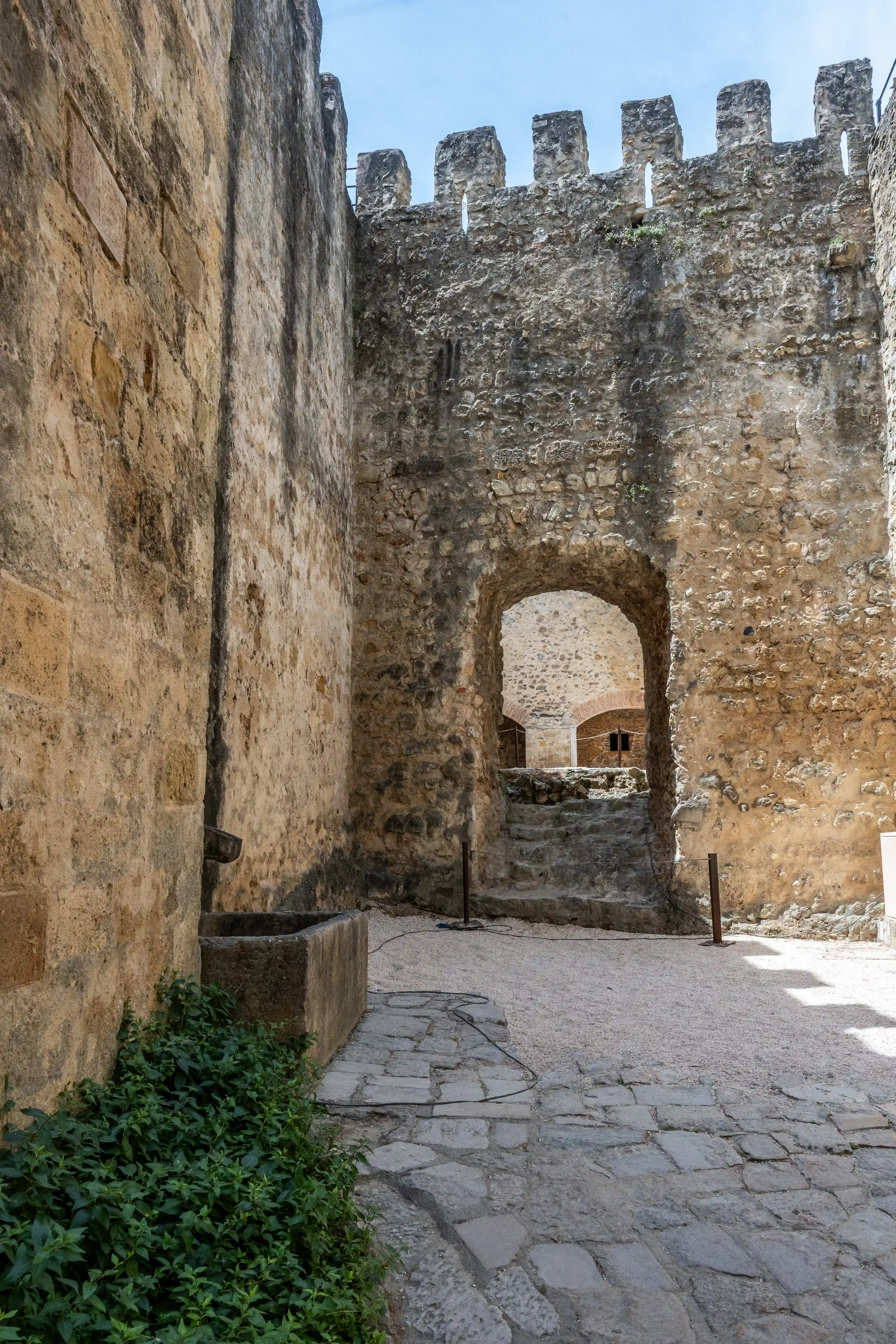 Arched stone gateway of an ancient castle courtyard, with weathered walls, a stone trough, green plants, and a sunlit paved area.