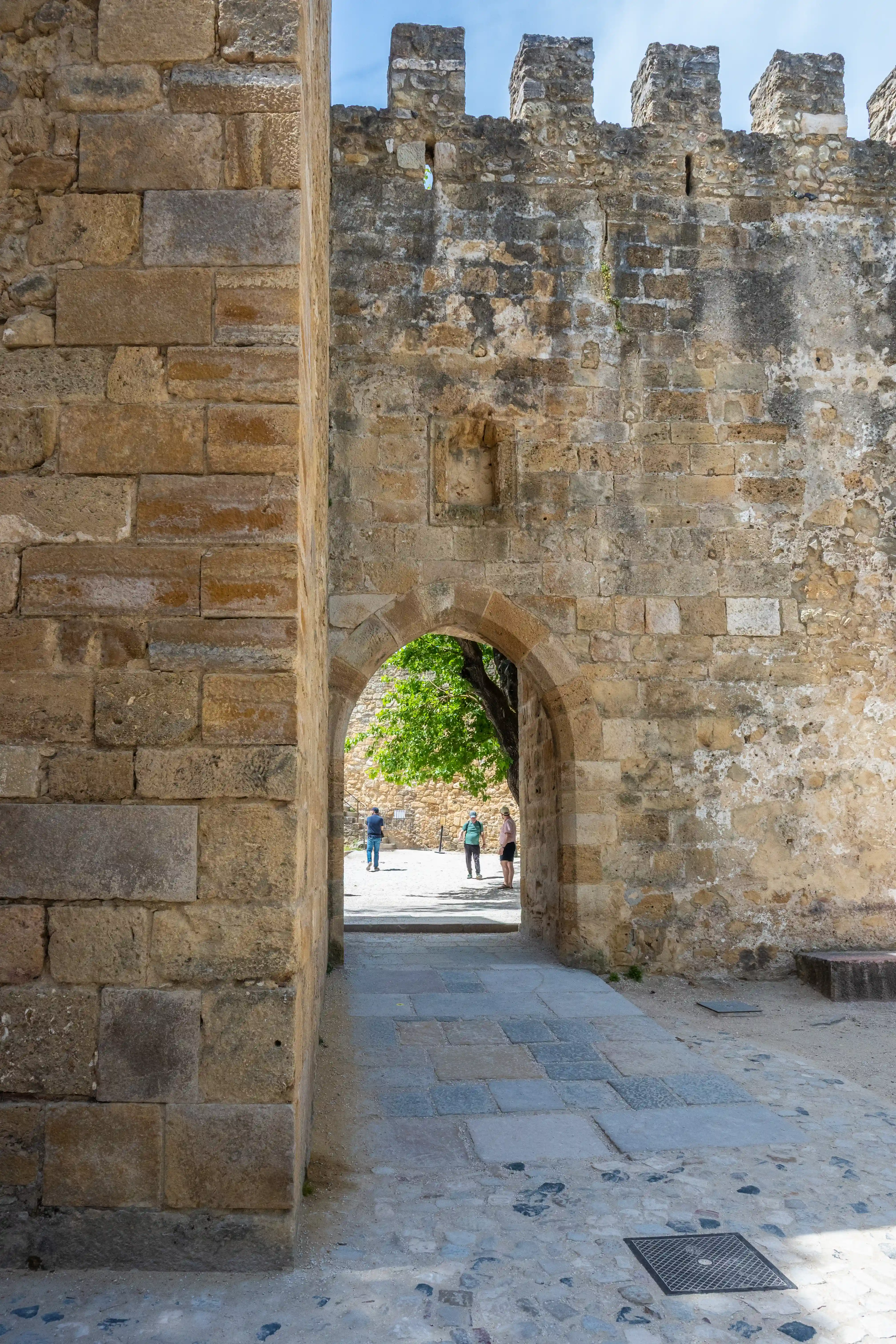 View through an ancient stone archway of a castle, with people walking in the sunlit courtyard beyond.