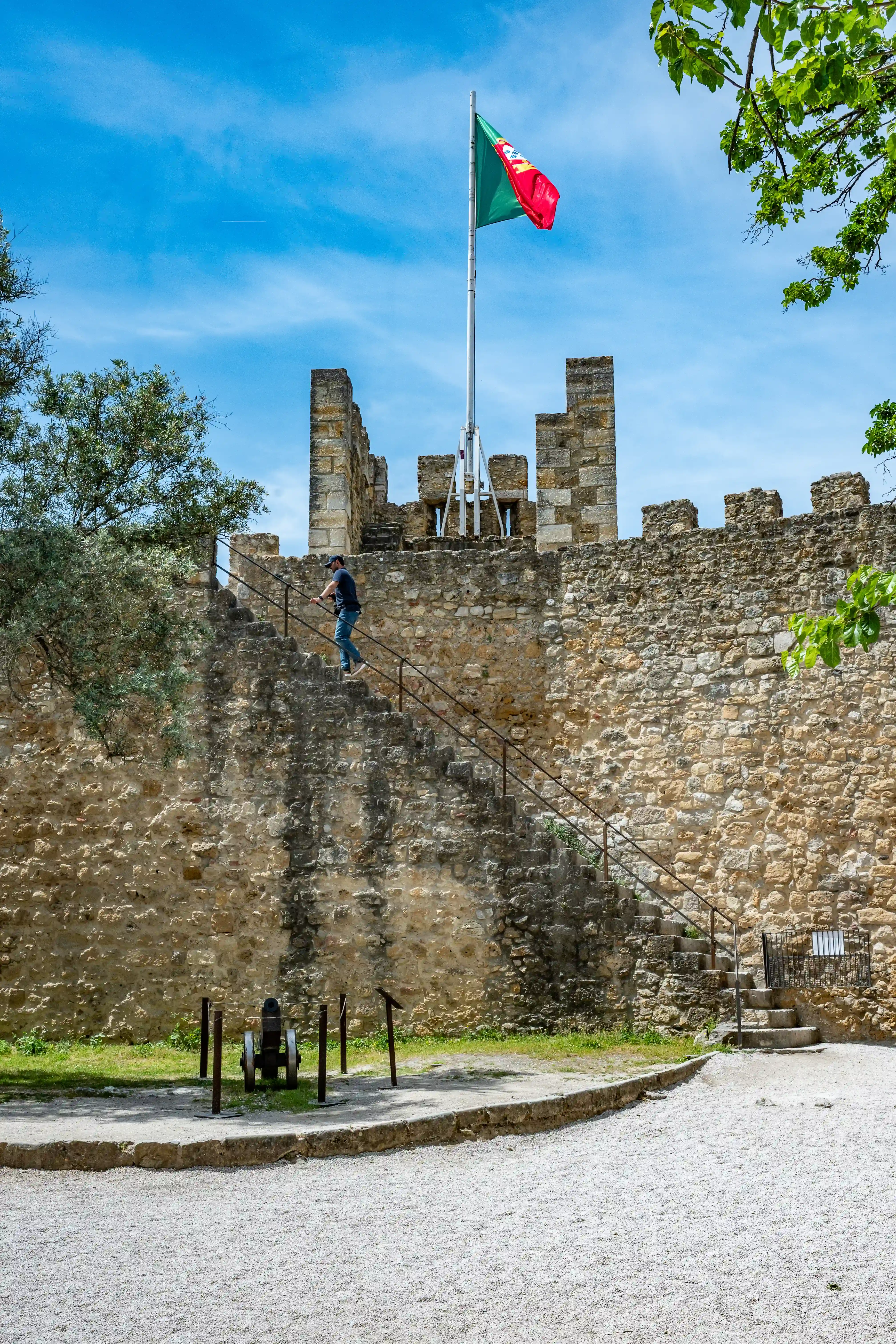 A person walks up the stairs of a stone castle wall with the Portuguese flag flying above. A small cannon is visible at the base of the wall.