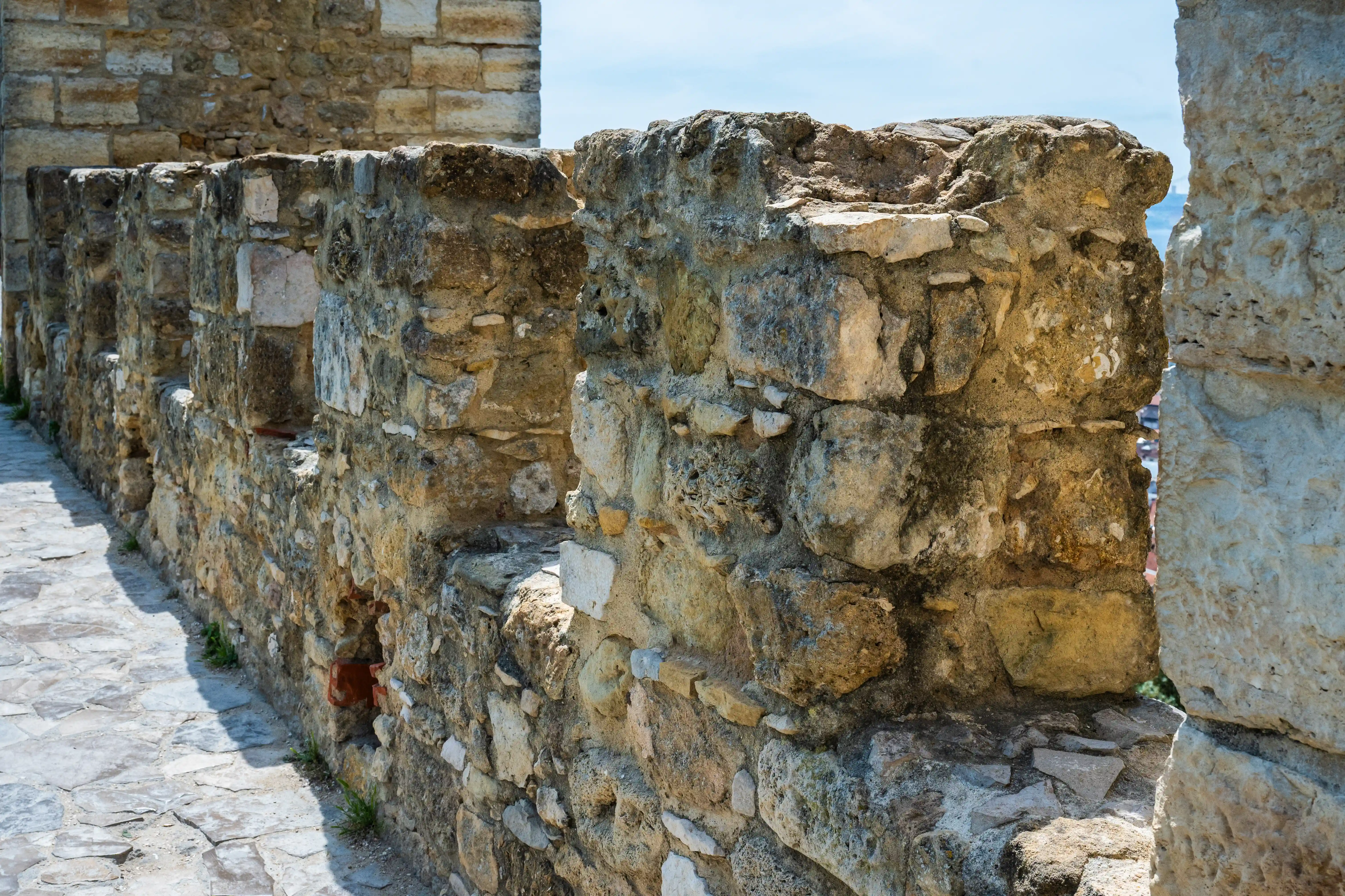 A close-up view of a rugged, ancient stone wall with several openings, casting shadows on a paved walkway below.