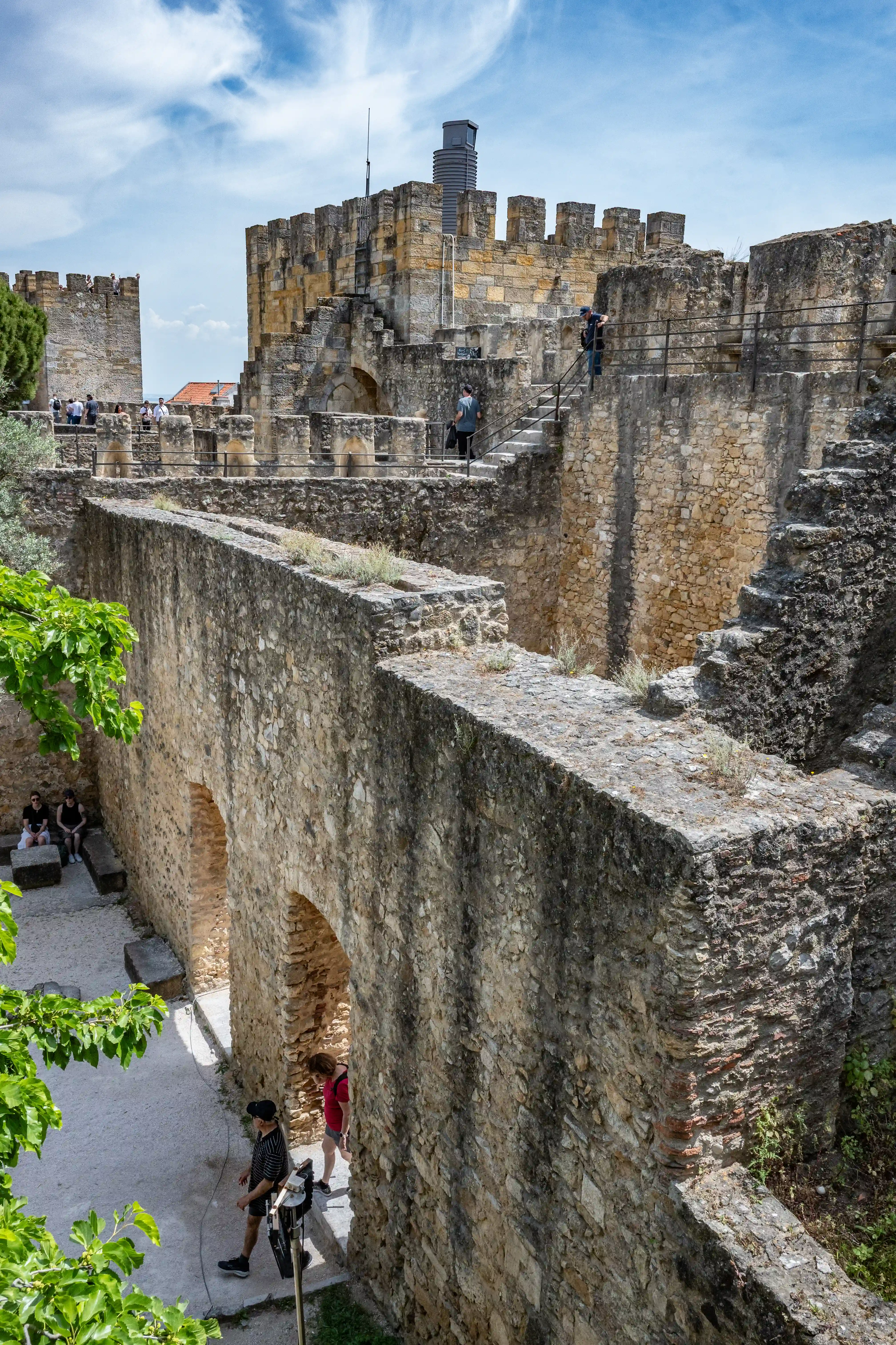 A stone castle with crenellated walls and towers under a partly cloudy sky. People are walking on the ramparts and stairs.