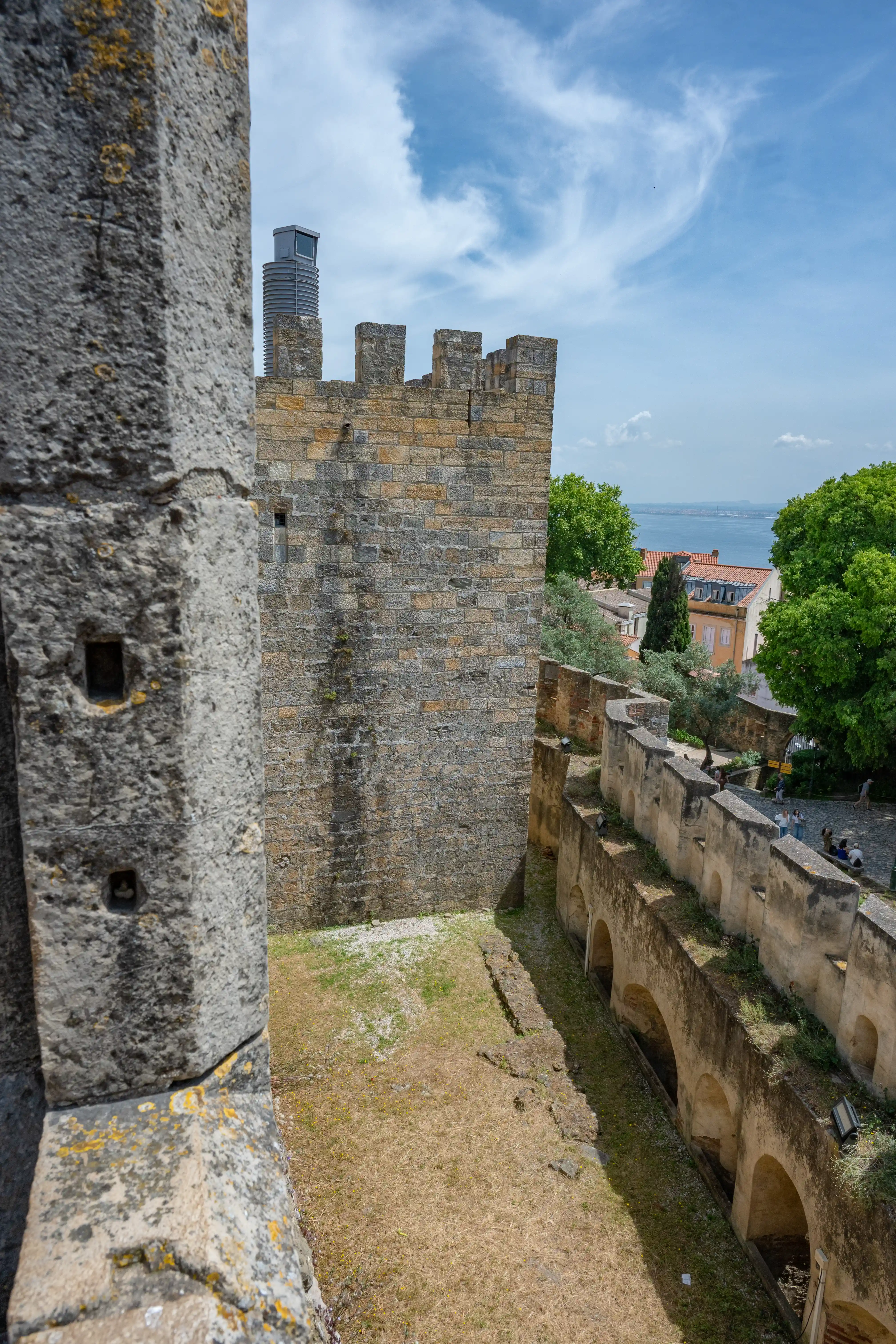A high-angle view from ancient stone castle battlements, looking down onto a dry courtyard and a wall with arched openings. In the background, a city with red-tiled roofs, trees, and the sea can be seen under a blue sky with white clouds.