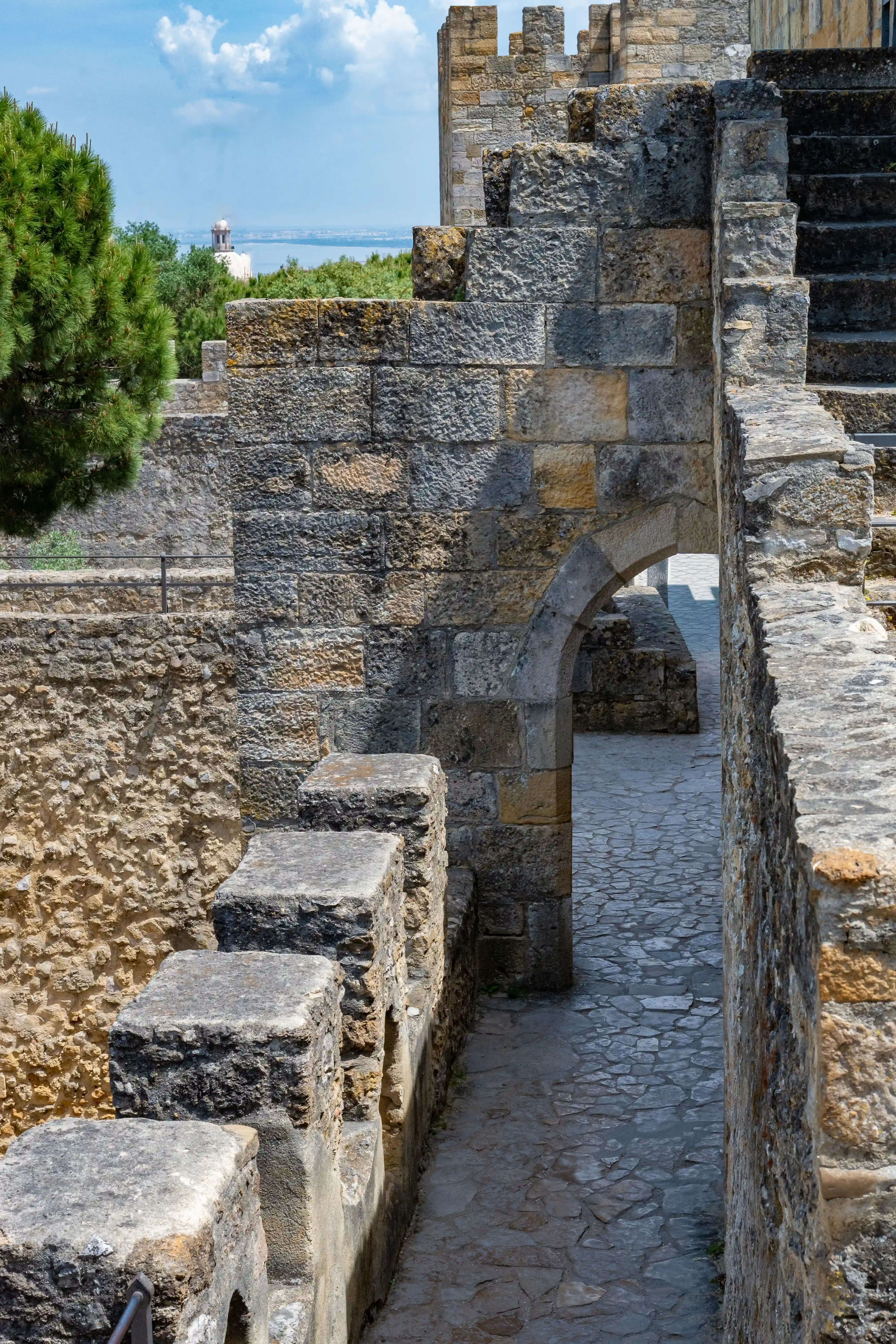 A sunlit scene inside a historic stone castle, showing a walkway bordered by thick, rough-hewn stone walls. A stone archway frames a view of the path beyond, where two people are visible. A large evergreen tree stands to the left, and in the far distance, a crane and a body of water are visible under a cloudy sky.