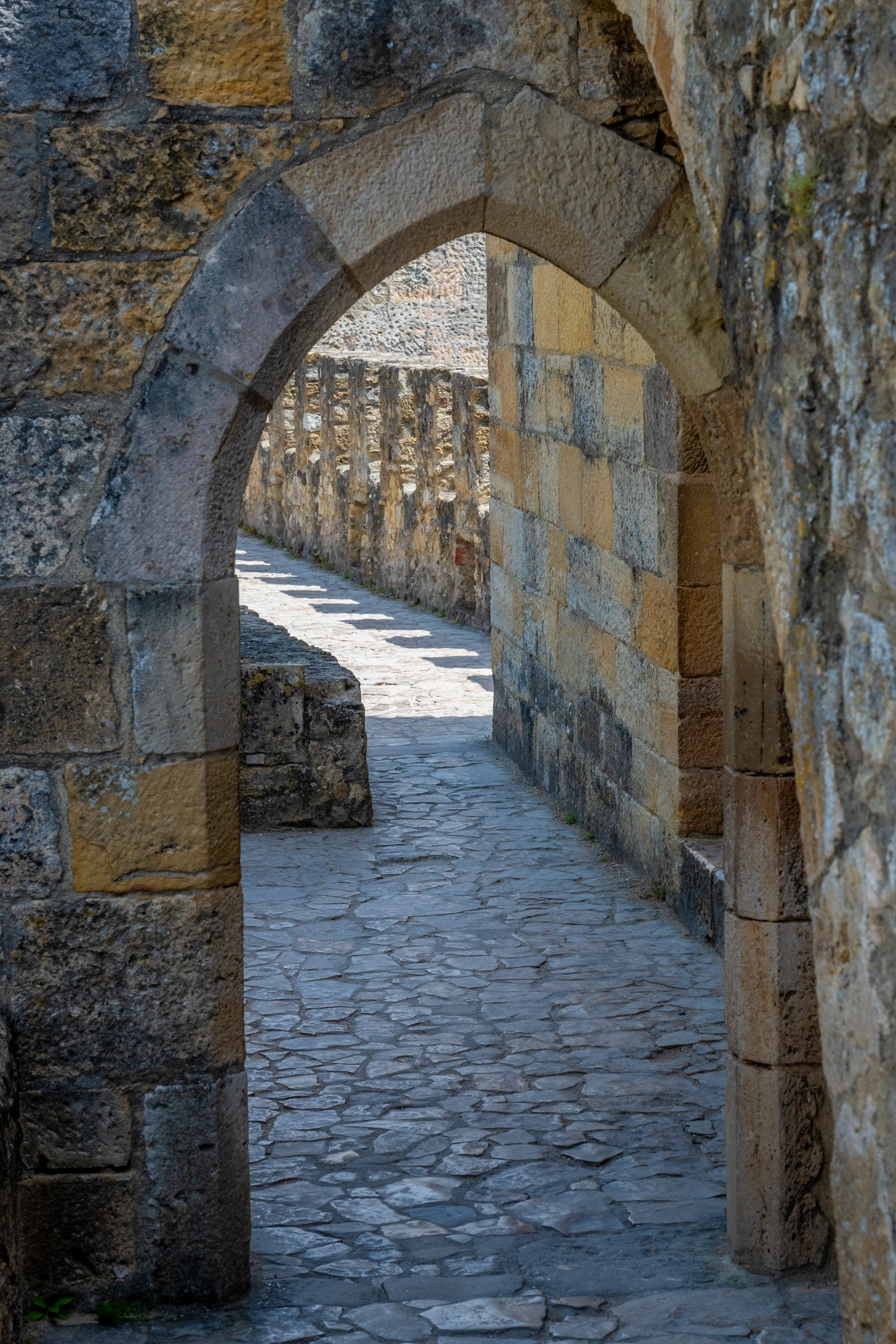 A stone archway frames a pathway within an ancient stone building, with sunlight and shadows on the cobblestone floor.
