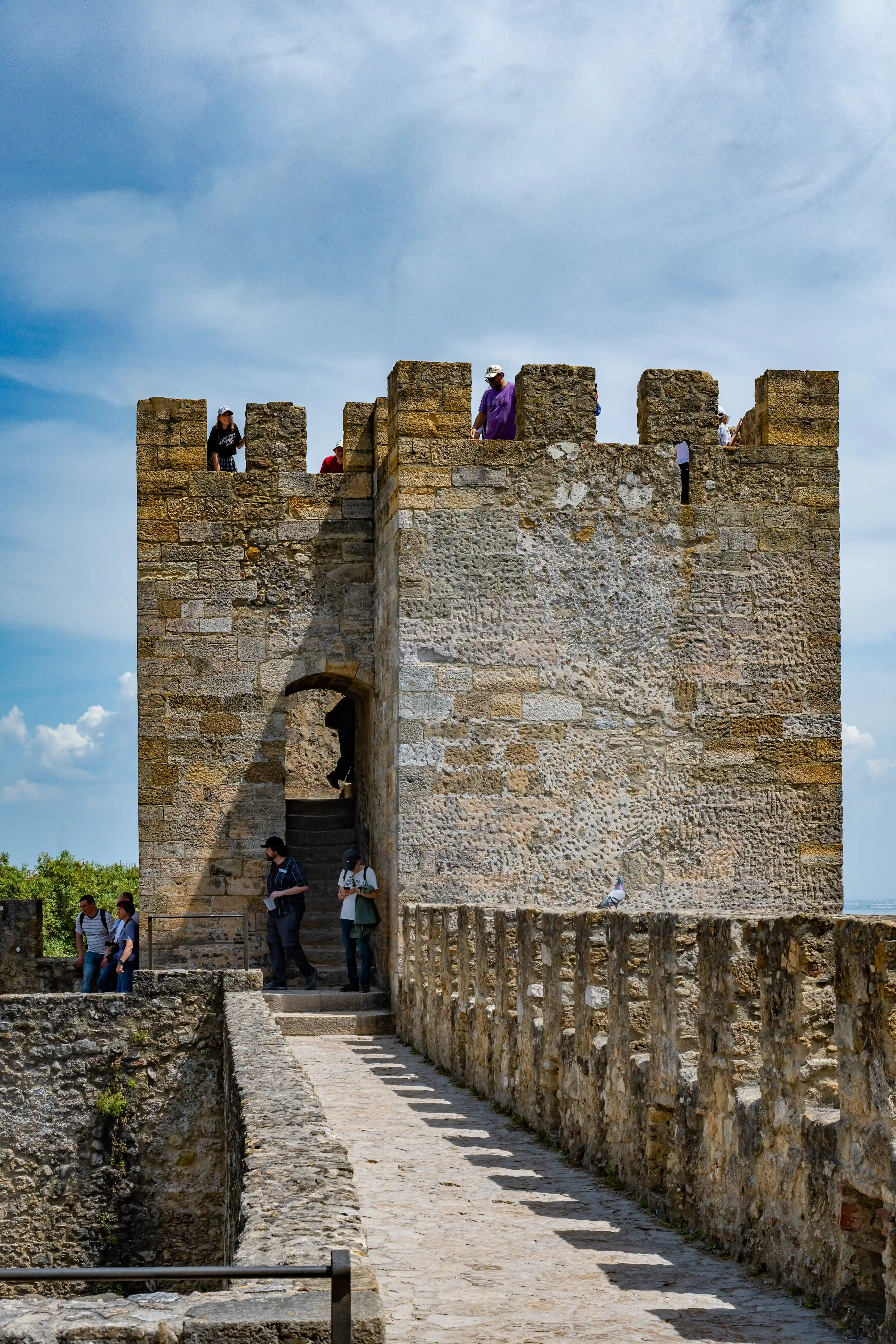 A group of people walk along a stone pathway on the ramparts of a castle. A tall stone tower with an arched entrance is visible. The sky is blue with some clouds.