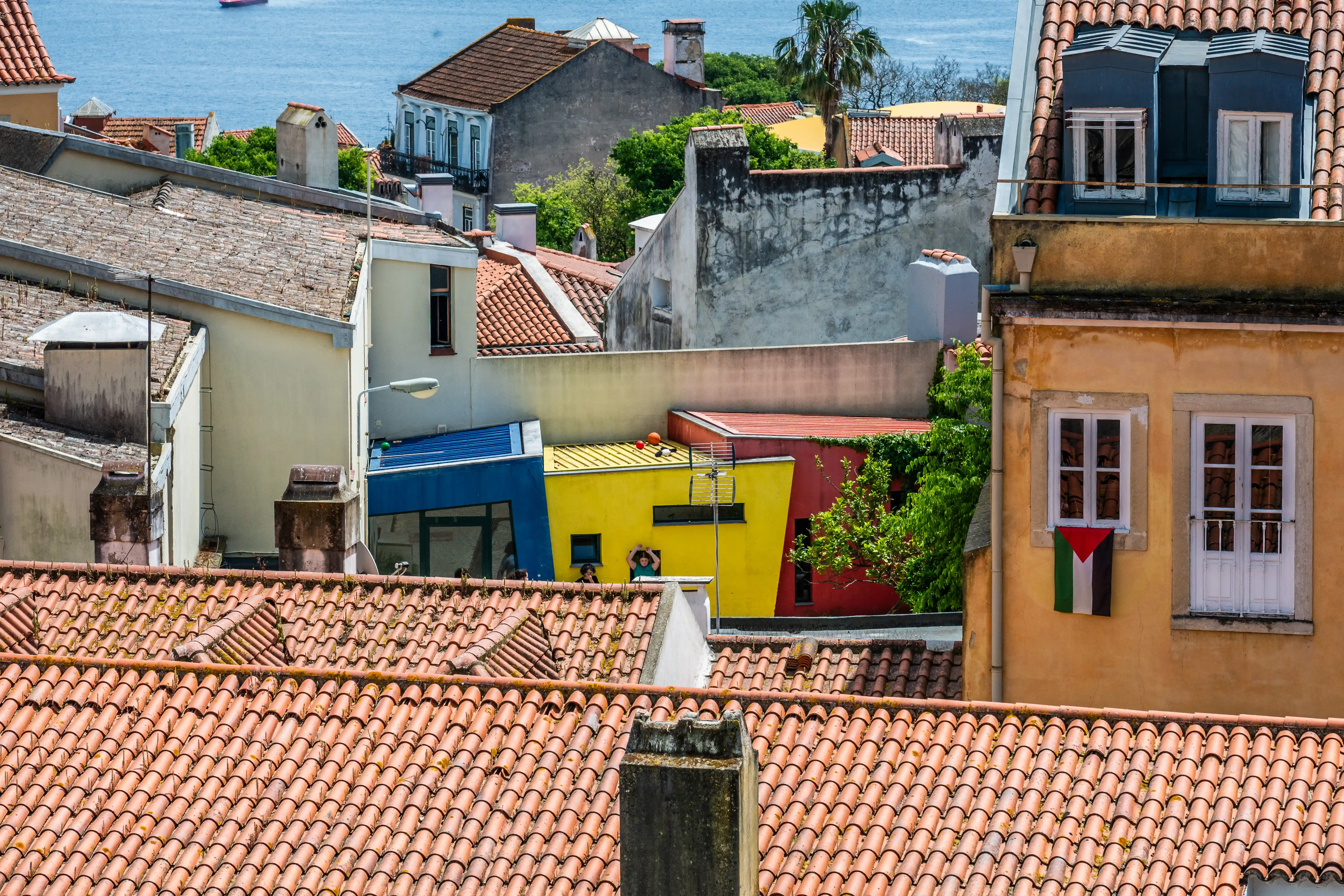 A view from above of a cityscape with many terracotta tiled roofs. In the middle ground, several buildings are painted in bright colours: blue, yellow, and red. A Palestinian flag hangs from a window on a yellow-ochre building on the right. The sea is visible in the background with a ferry boat.