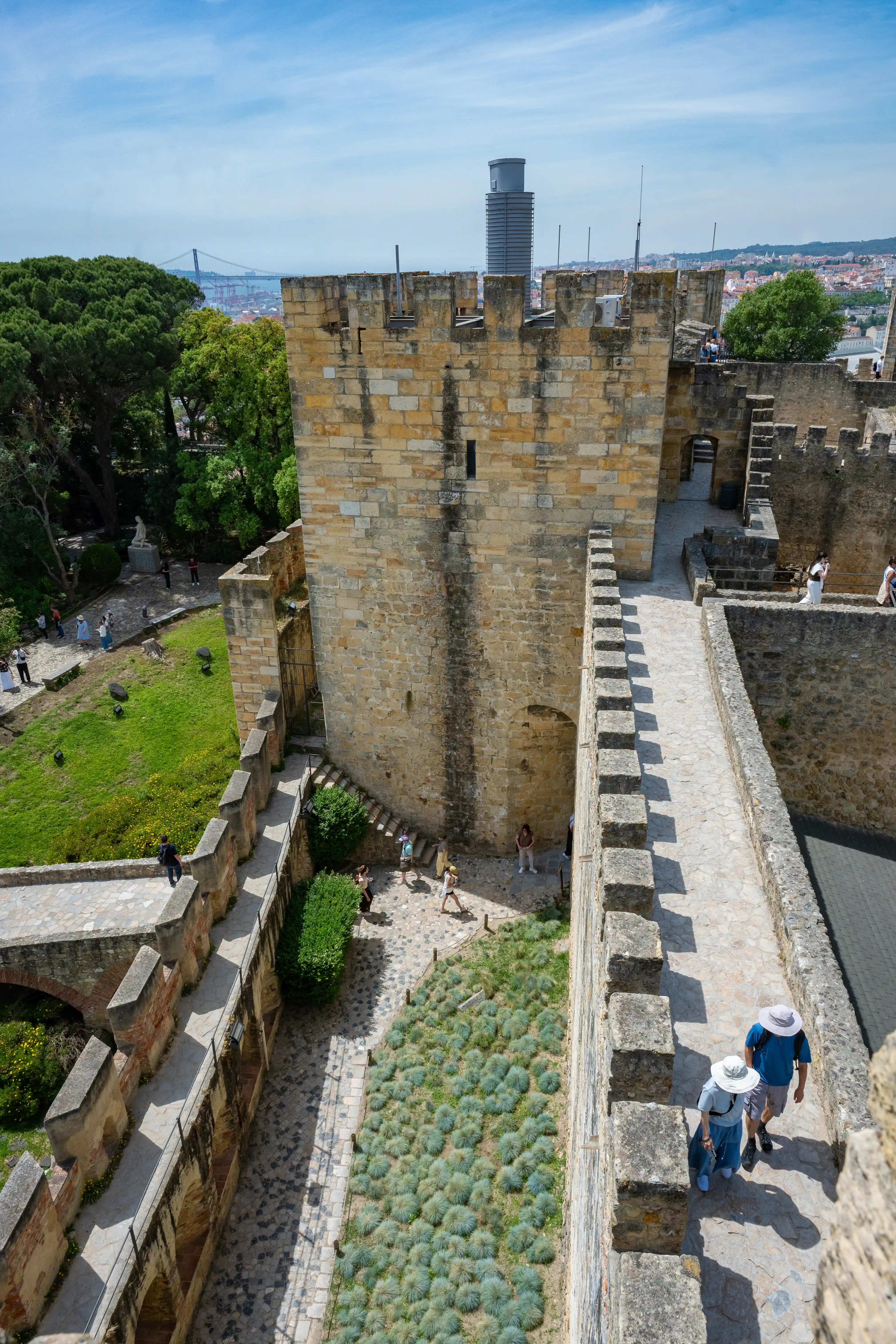  People walking on the stone ramparts of an old castle, with trees, greenery, and the city skyline in the background.