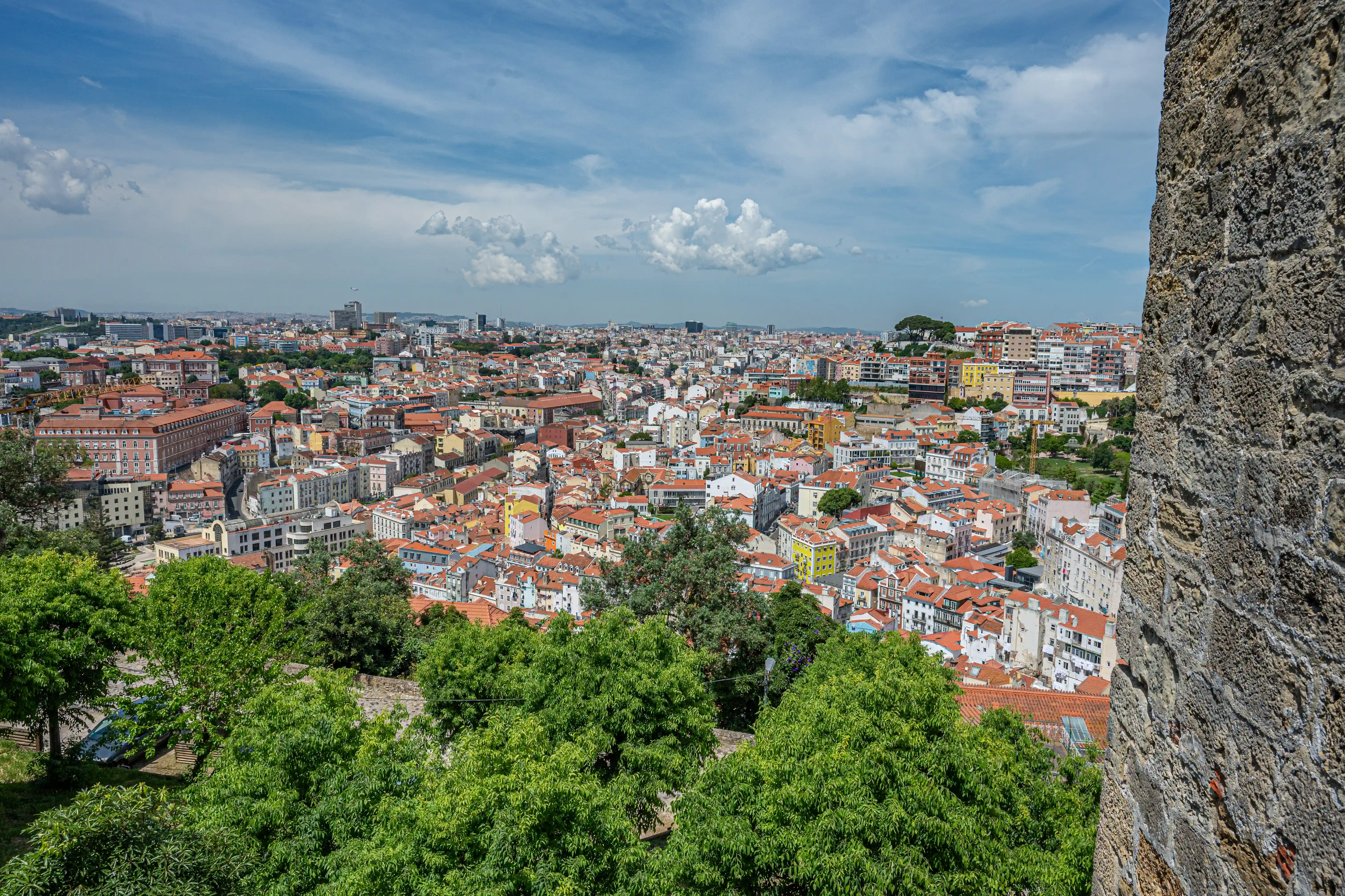 View over the rooftops of Lisbon, Portugal, with terracotta tiles and white buildings stretching to the horizon under a sky with fluffy white clouds. A textured stone wall is in the foreground.